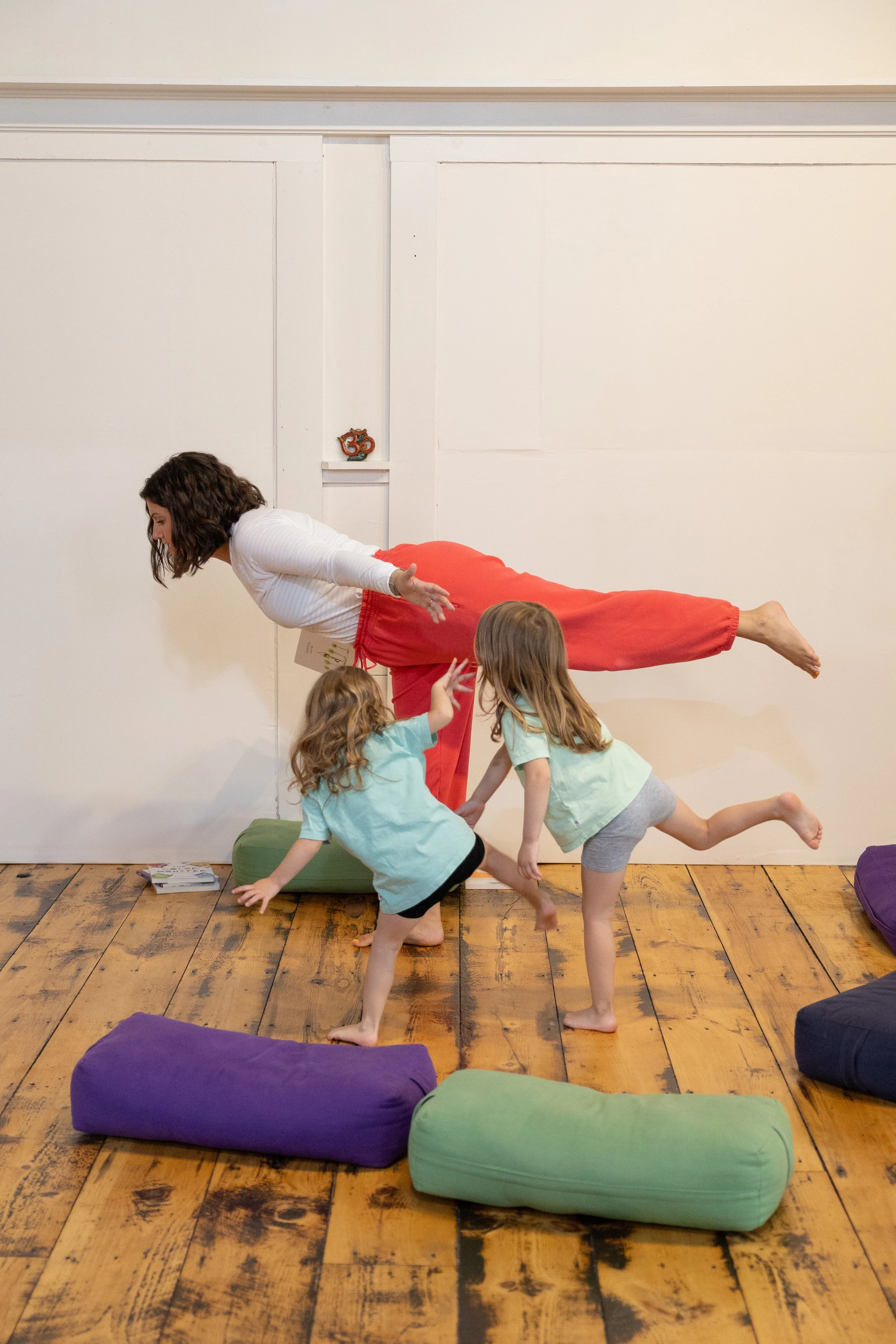 Two young girls and an adult woman are practicing yoga in a room with wooden floors and white walls. The woman is balancing on one foot with her other leg extended behind her, while the girls are reaching out to her, assisting or mimicking her pose. Colorful yoga bolsters are on the floor.