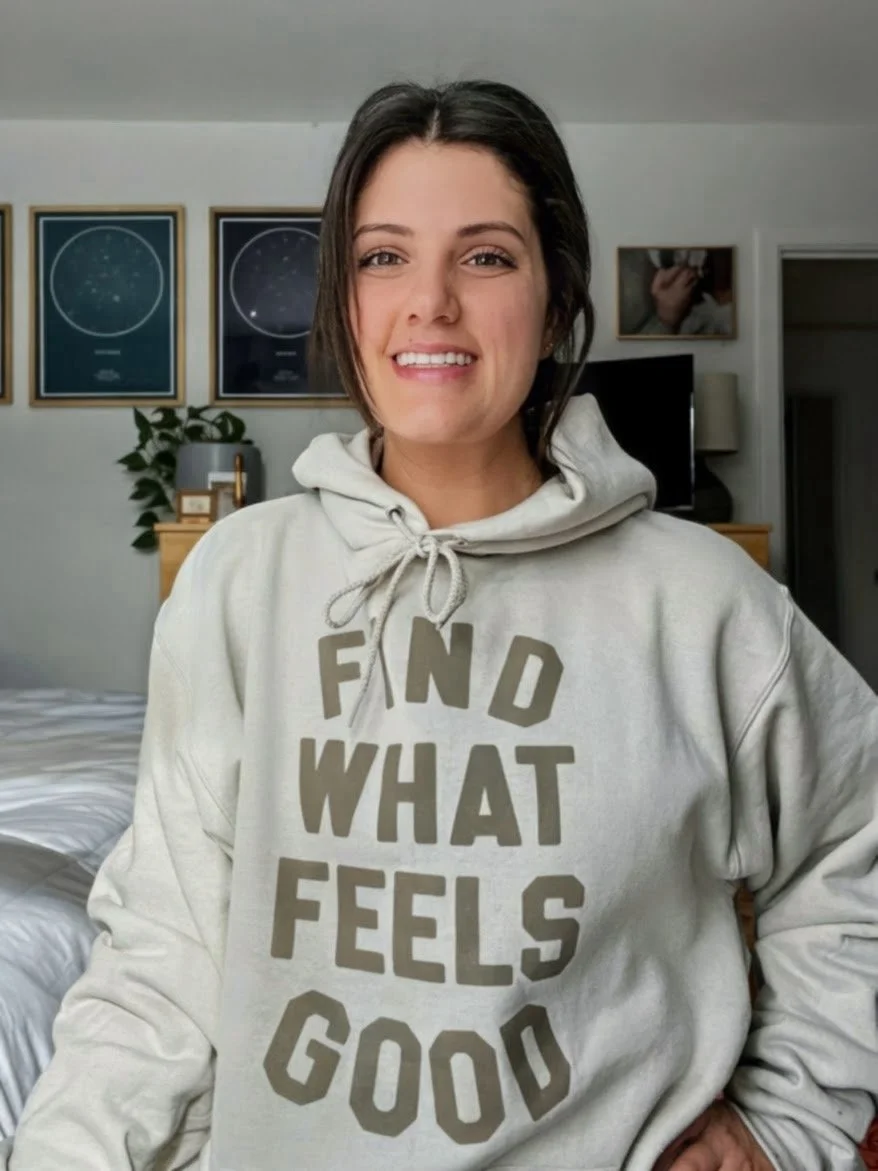 A smiling woman with shoulder-length dark hair wearing a gray hoodie with the words 'FIND WHAT FEELS GOOD' in large letters, standing in a bedroom.