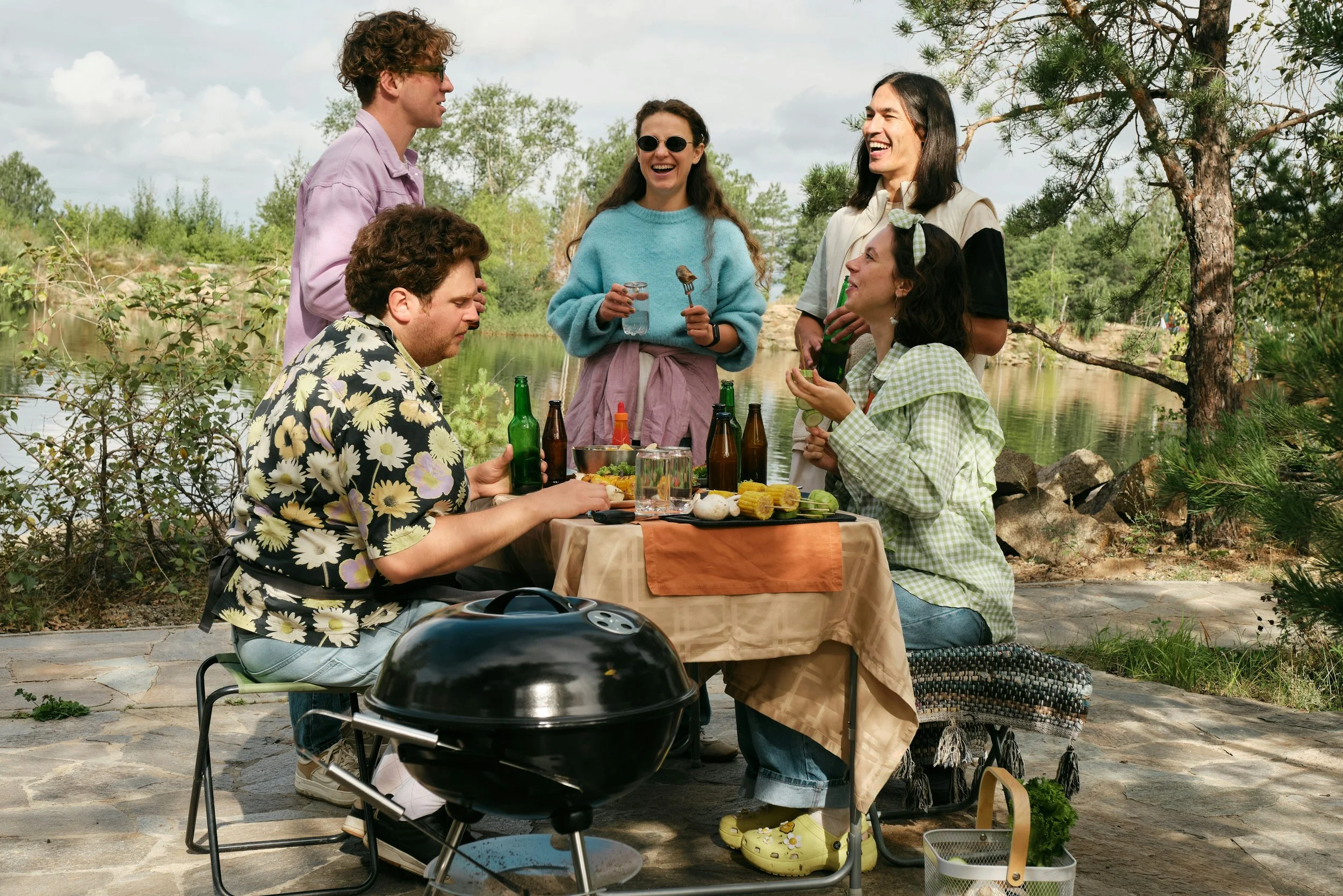 a group of 5 friends at a picnic by the river. a black bbq is in the foreground, and there is a small folding table they are sitting and standing around