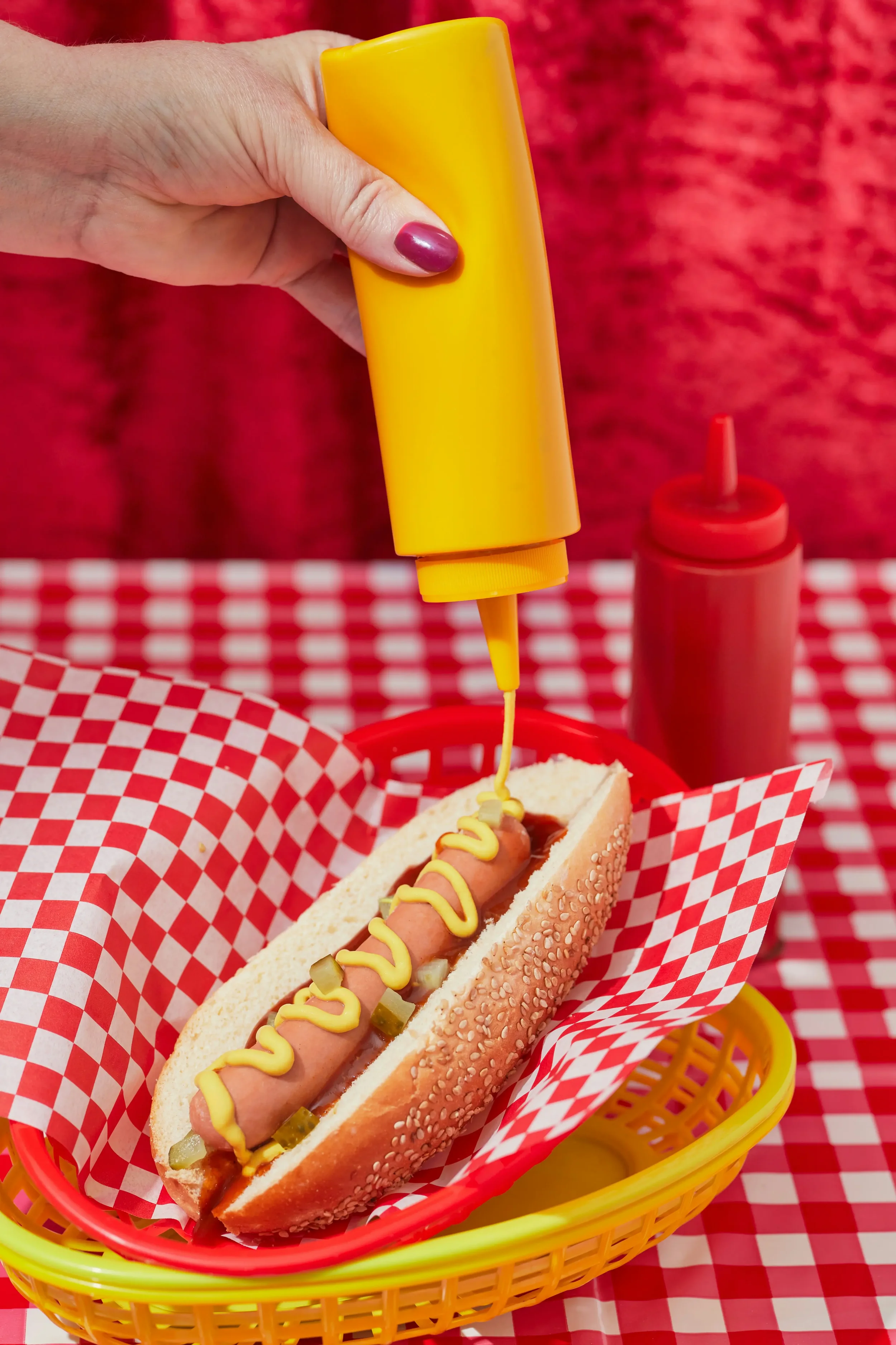 A yellow bottle of mustard sauce being squirted onto a hot dog in a bun with a red and white checked background