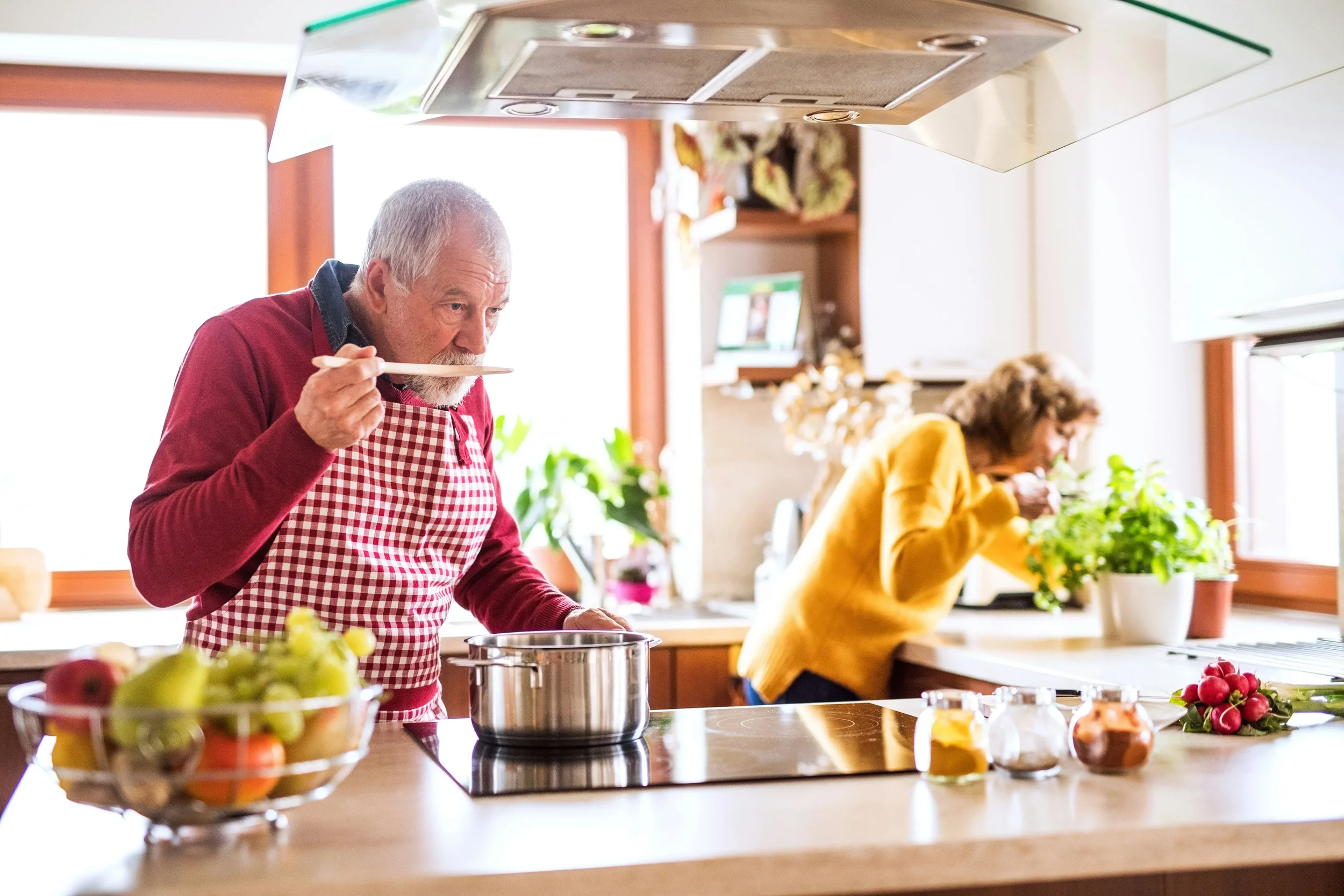 a man and a woman in a cozy home kitchen. The man is wearing a red jumper with a red and white check apron. the woman is wearing a mustard coloured jumper. She is picking some fresh herbs from a pot on the counter. He is tasting a dish he is cooking.