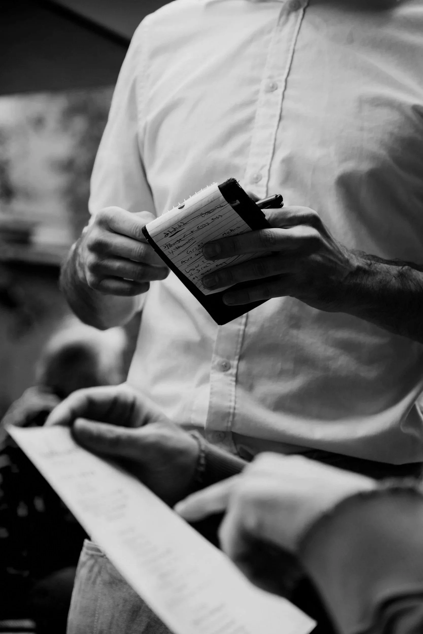 black and white image of a waiter taking an order on a little pad with a pen. The customers hand and menu can be seen in the foreground
