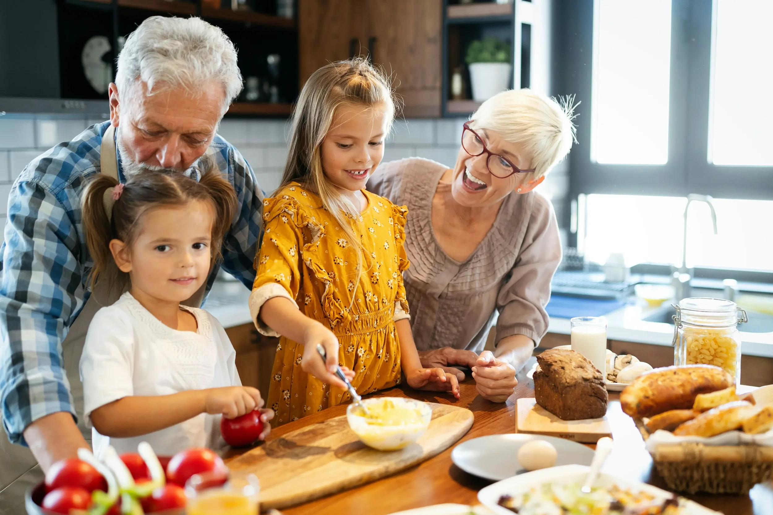 a multigenerational family in a cozy home kitchen. There are two older people (man and woman) and two little girls. they are cooking a dish together.