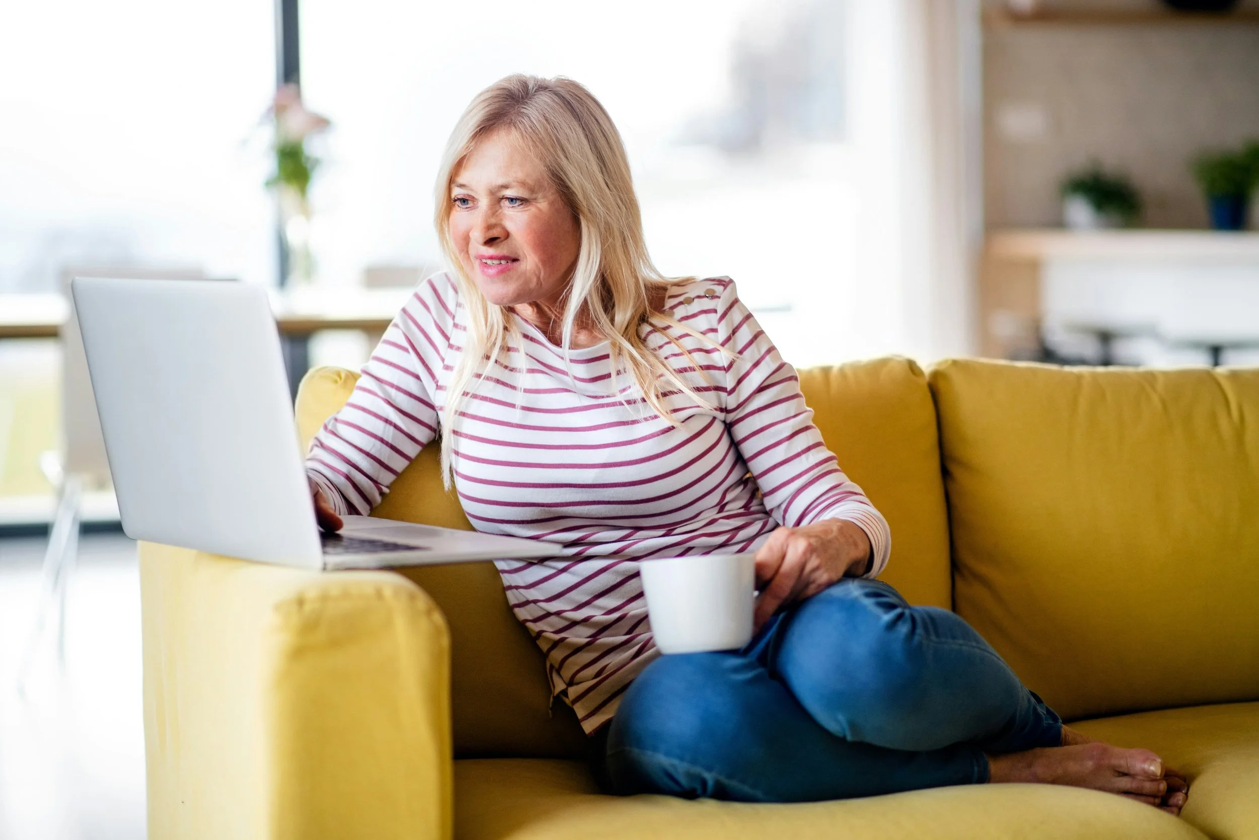 caucasian woman in striped red top with blue jeans sitting on a mustard couch looking at a laptop with a cup of coffee in her hand.