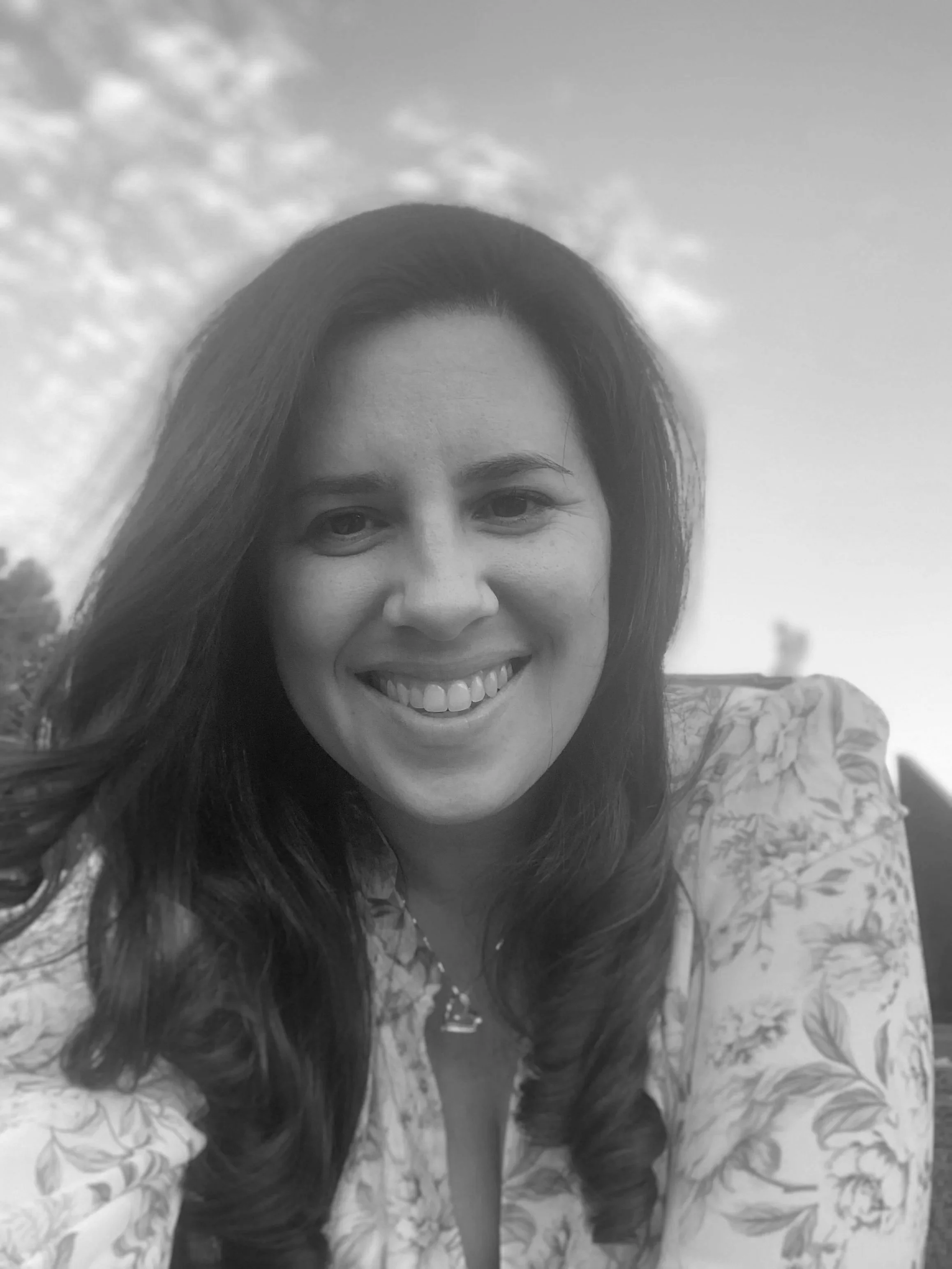 Black and white headshot of Elyse, who has long brown hair and is wearing a floral dress. She is smiling.