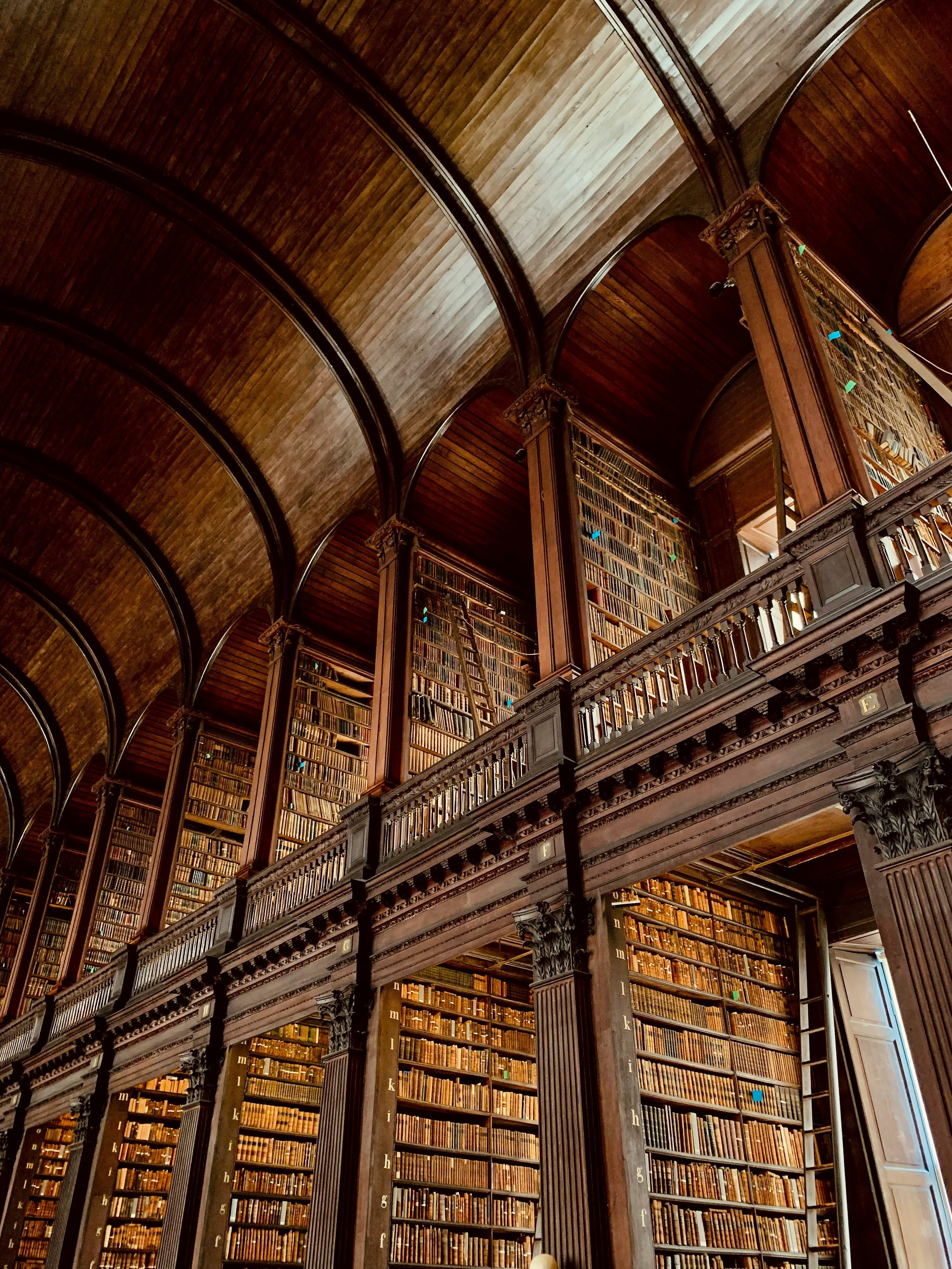 Interior of a historic library with tall wooden bookshelves filled with books, arched wooden ceiling, and ornate balcony with columns and railings.