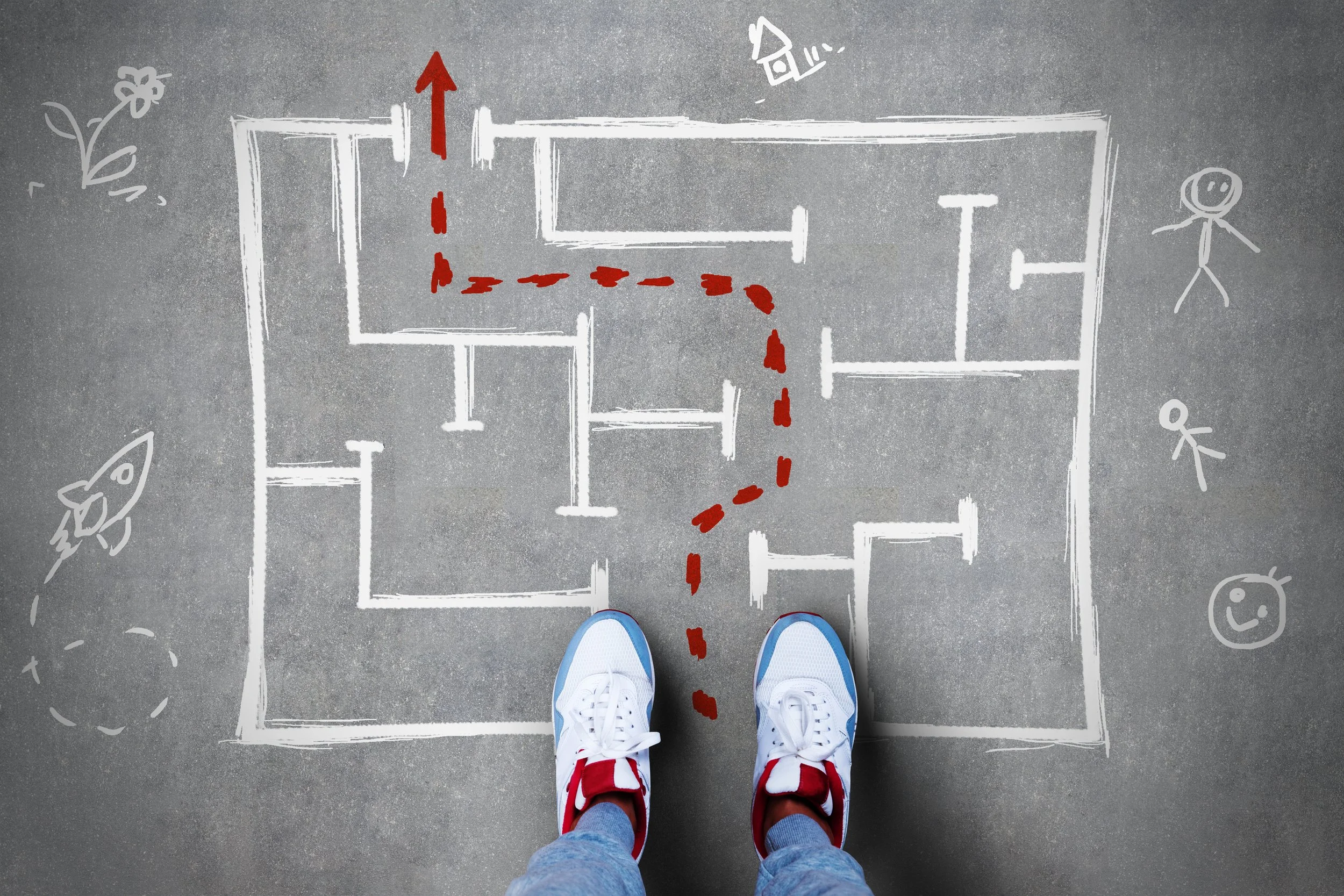 A person wearing white sneakers and jeans standing next to a chalk-drawn floor plan on a gray pavement. The floor plan shows rooms and red dashed lines indicating movement paths. Drawings of a flower, rocket, ice cream cone, and smiling face are around the plan.