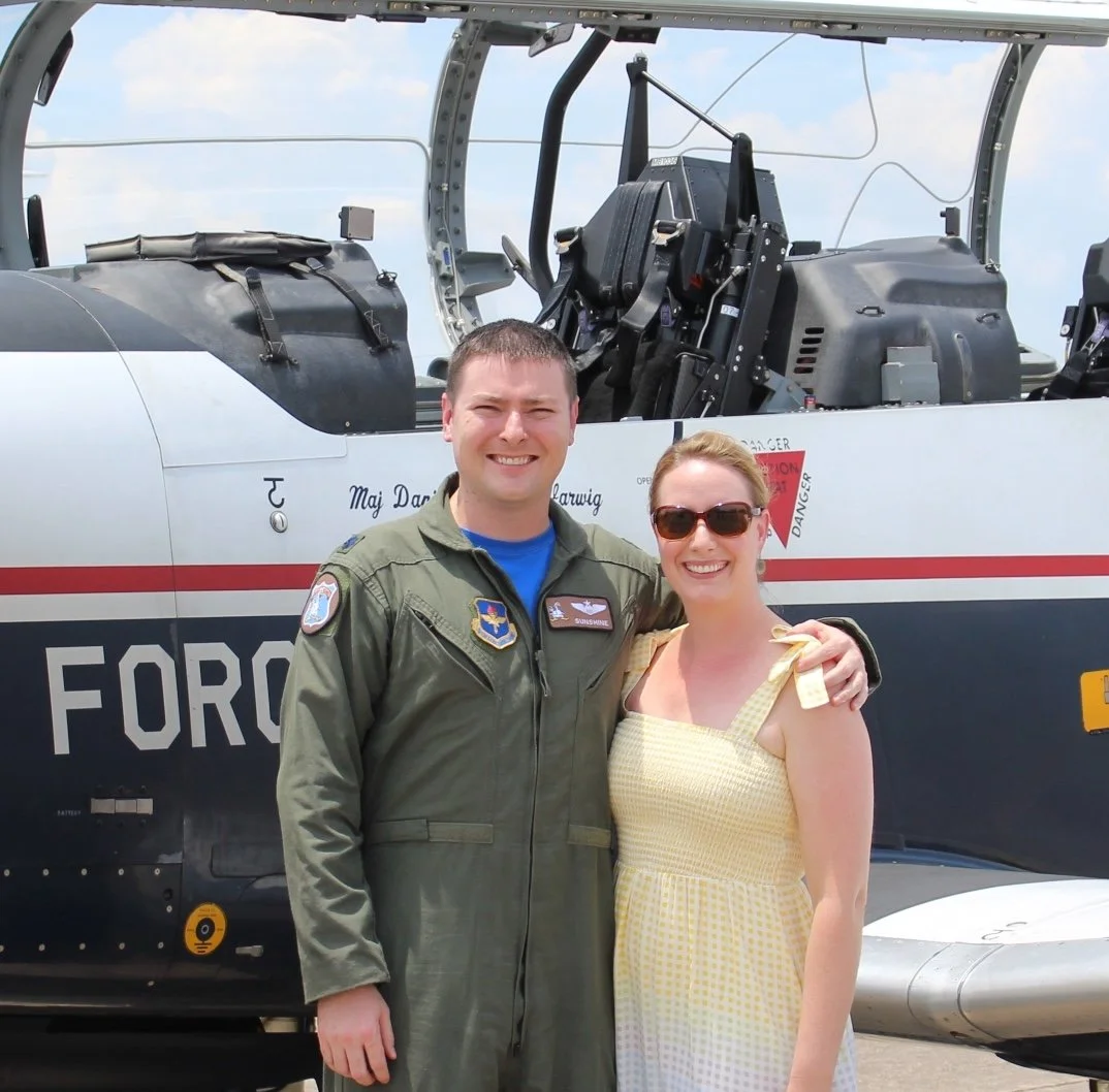 A man in military flight suit and a woman in a yellow dress smiling and standing in front of a fighter jet.