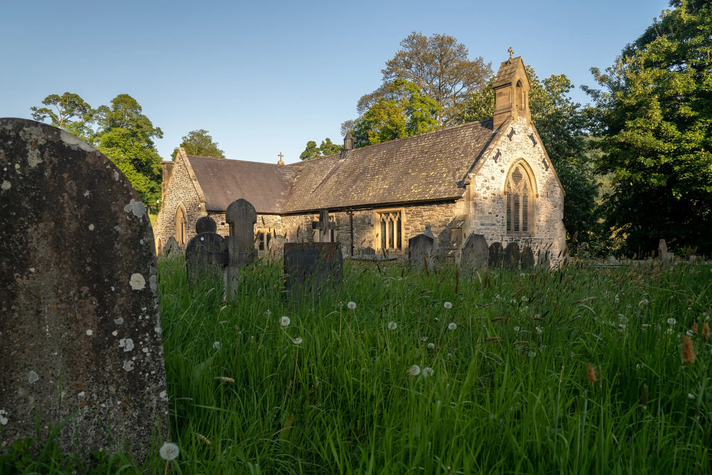 A small stone church with a steeple and arched windows, surrounded by a graveyard with age-weathered gravestones, lush green grass, and tall trees in the background under a clear blue sky.
