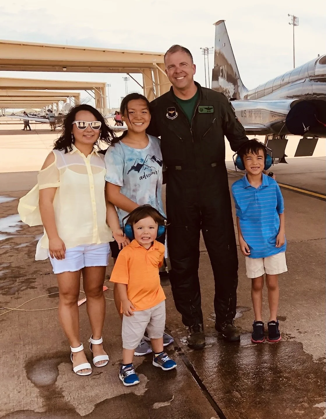 A group of five children and one adult standing in front of a jet airplane at an airfield, smiling at the camera.