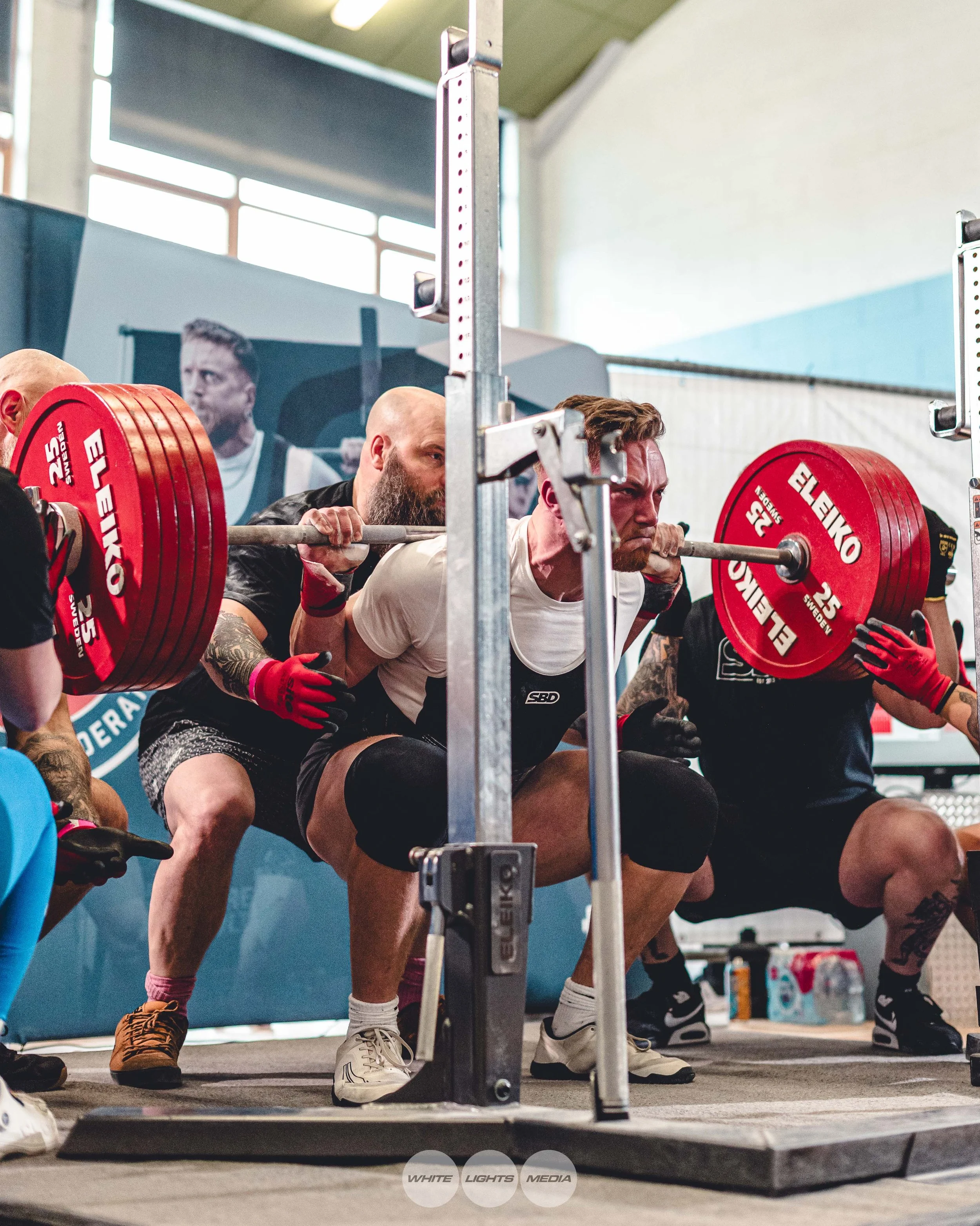 A man doing a squat with a heavily loaded barbell at a competition. Five spotters are assisting him, one directly behind and others to the sides.