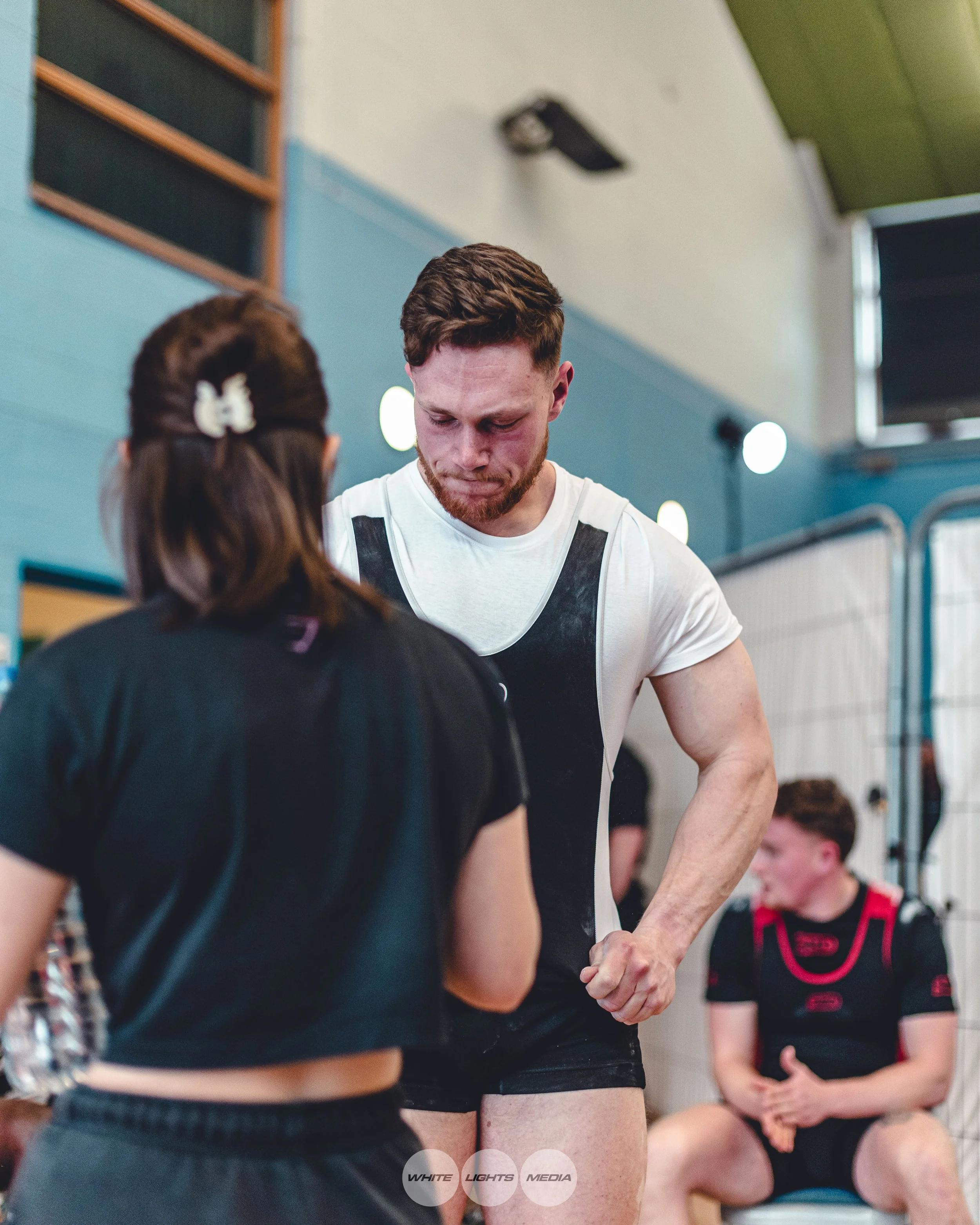 A man with a beard and short hair flexing his arm muscles while a woman with brown hair and a white butterfly hair clip talks to him, in a competition with other people in the background.
