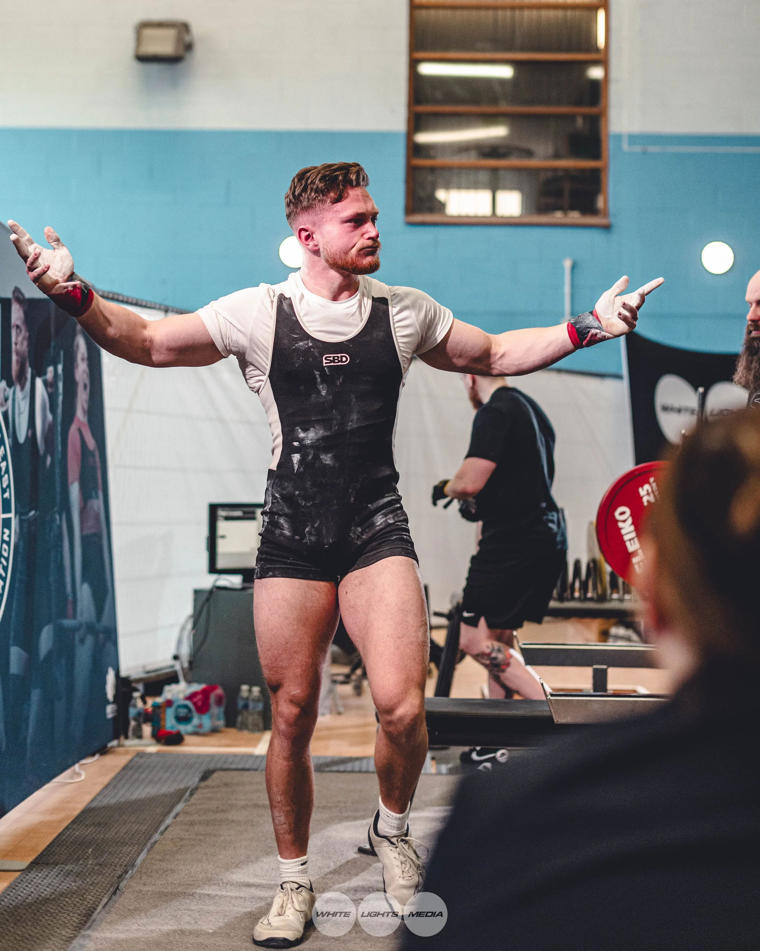 A male powerlifter wearing a black and white singlet, white t-shirt, and barefoot shoes, with chalk on his hands and body, standing with arms outstretched during a competition after completing his lift.