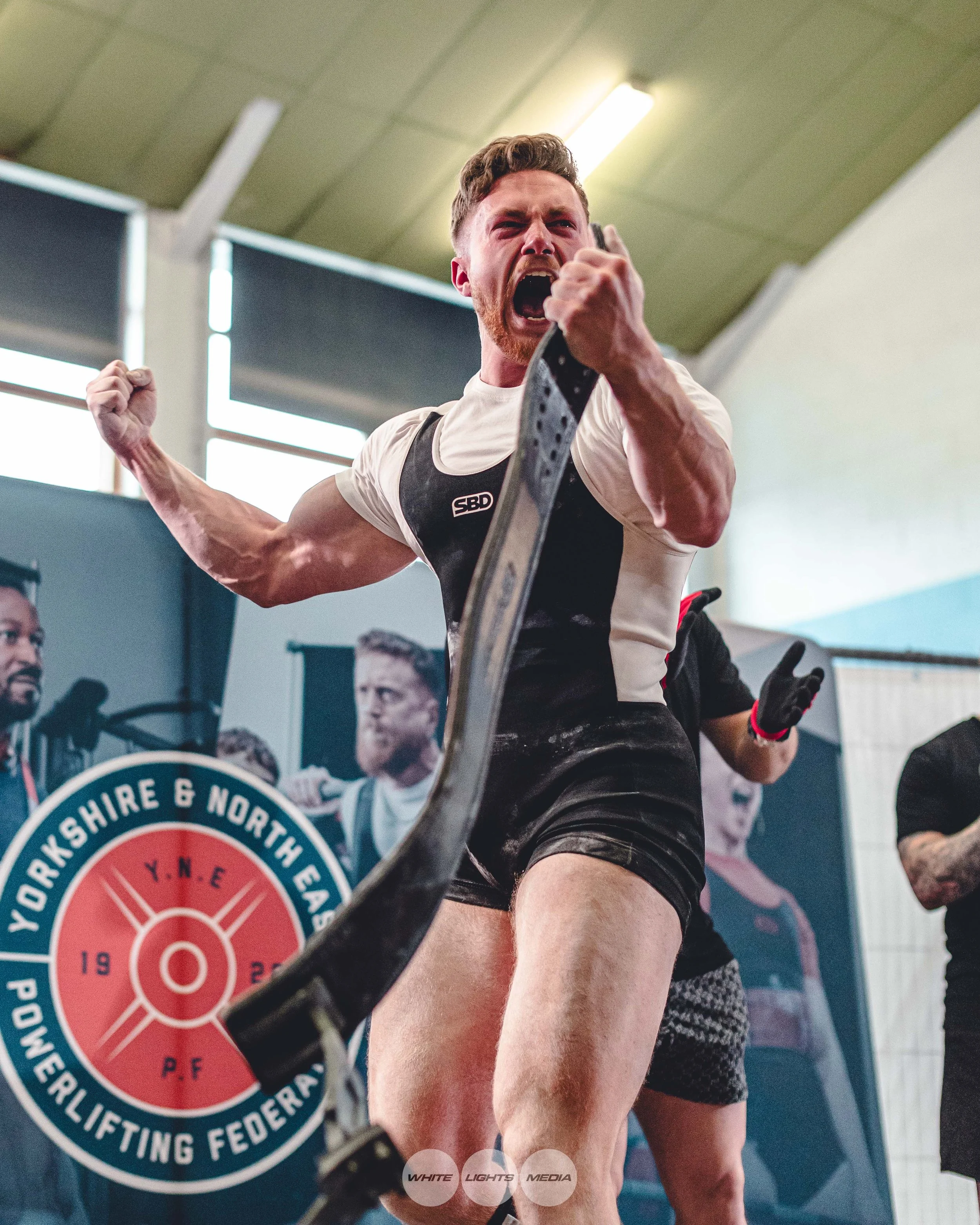 A muscular man shouting passionately while holding his belt during a powerlifting event.