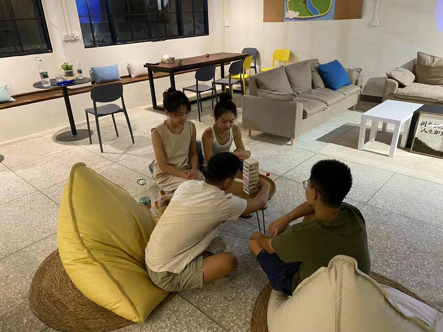 Four young people playing a game of Jenga on a small table in a cozy indoor space with couches and chairs.