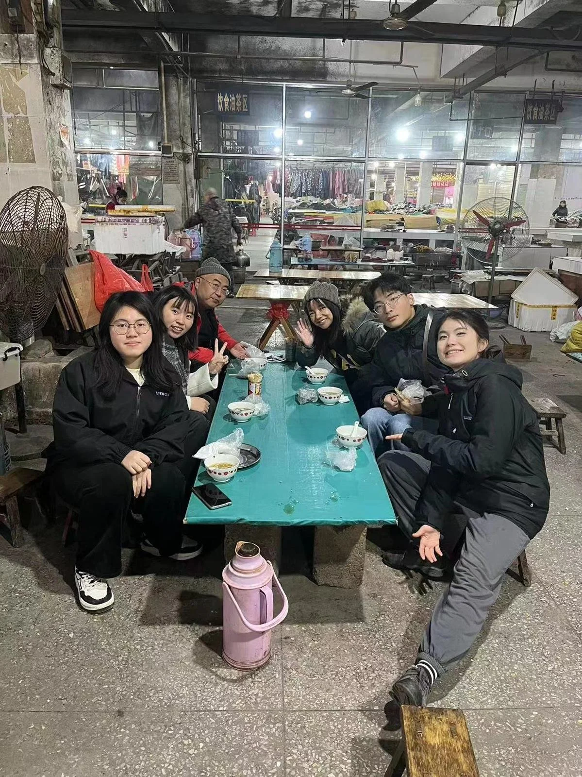 Six friends sitting around a table in an indoor market or food court, eating and smiling, with bowls and drinks in front of them, and market stalls selling clothes in the background.