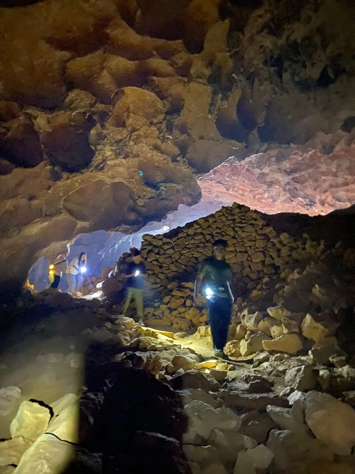 Group of people exploring a dark cave with headlamps and flashlights, surrounded by large rocks and uneven terrain.