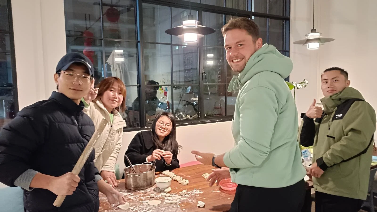 Five people in a kitchen making dumplings at a wooden table, smiling, with ingredients and cooking tools around, in a well-lit room with large windows.