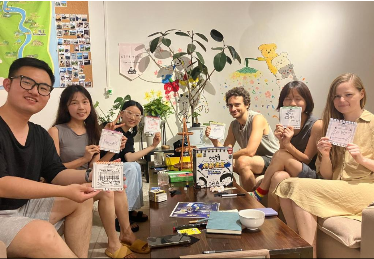 Group of seven young adults sitting around a wooden table in a cozy, decorated room, each holding small notebooks or sketches. The table has various items including books, papers, a bowl, and a small game box. The background features a colorful mural