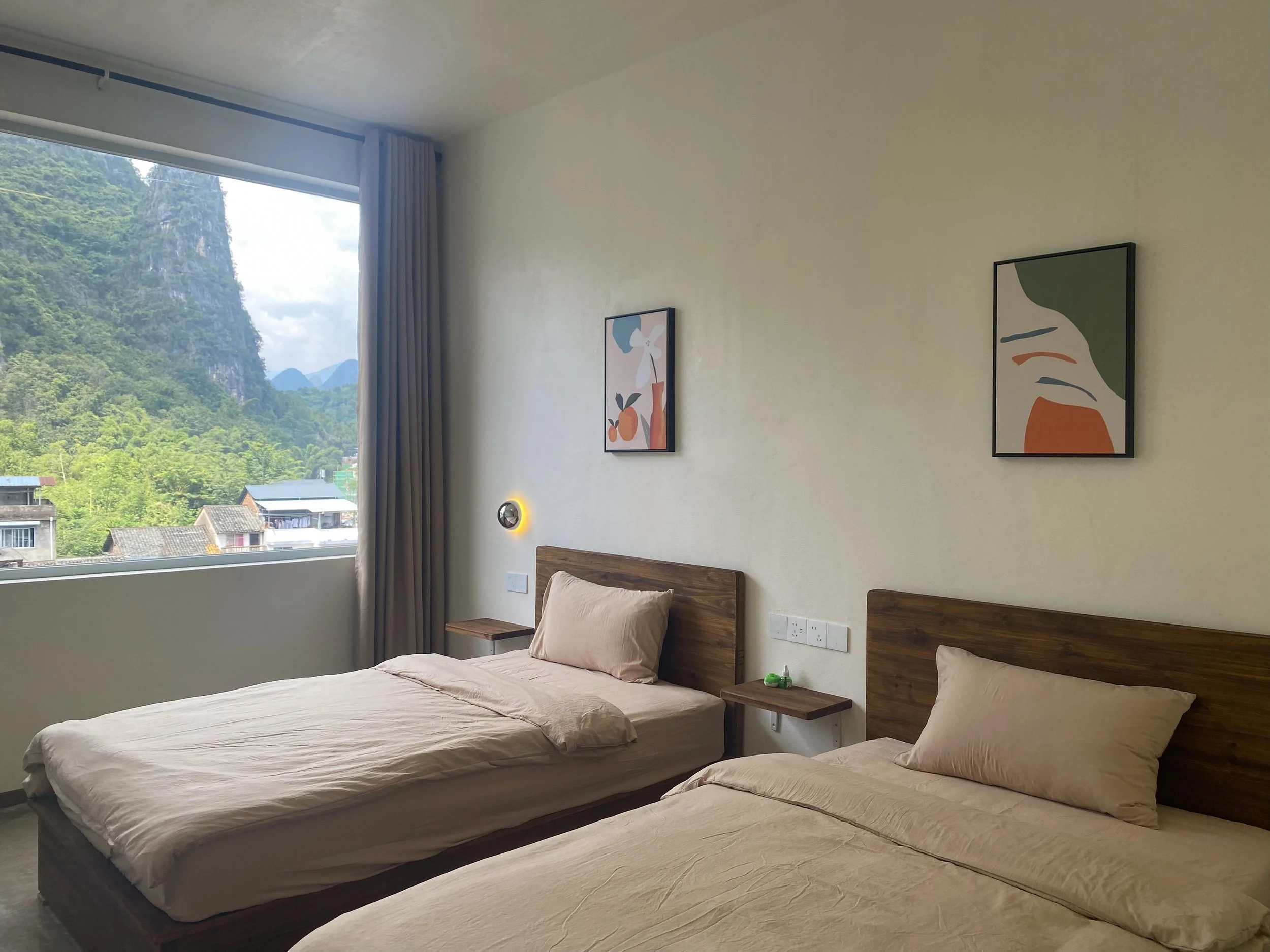 Hotel room with two beds, wooden headboards, beige bedding, small bedside tables, a window showing a view of green mountains and blue sky, and two abstract art paintings on the wall.
