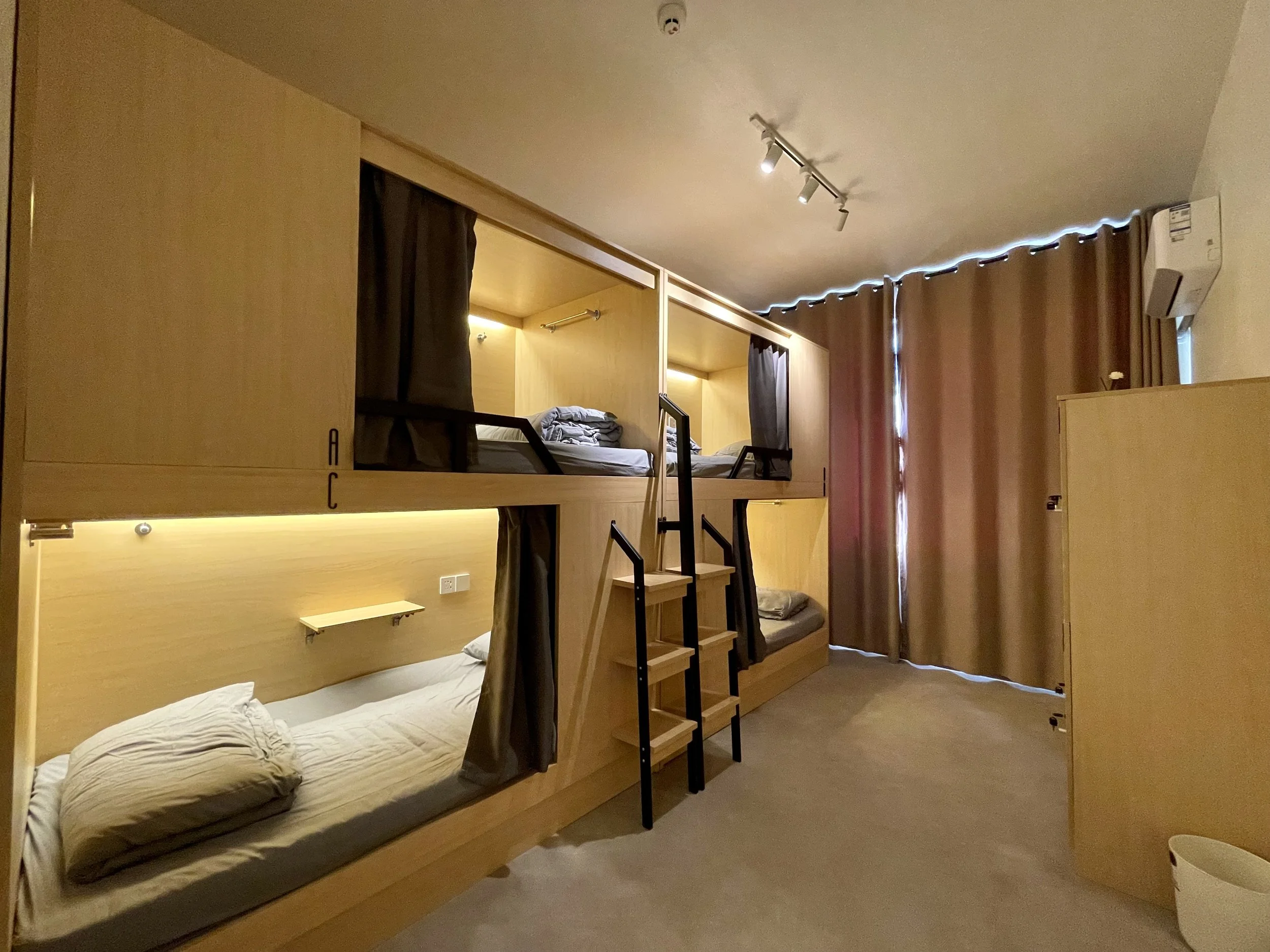 Room with wooden bunk beds and curtains in a hostel or capsule hotel, with stairs leading to the upper bunks and an air conditioning unit on the wall.