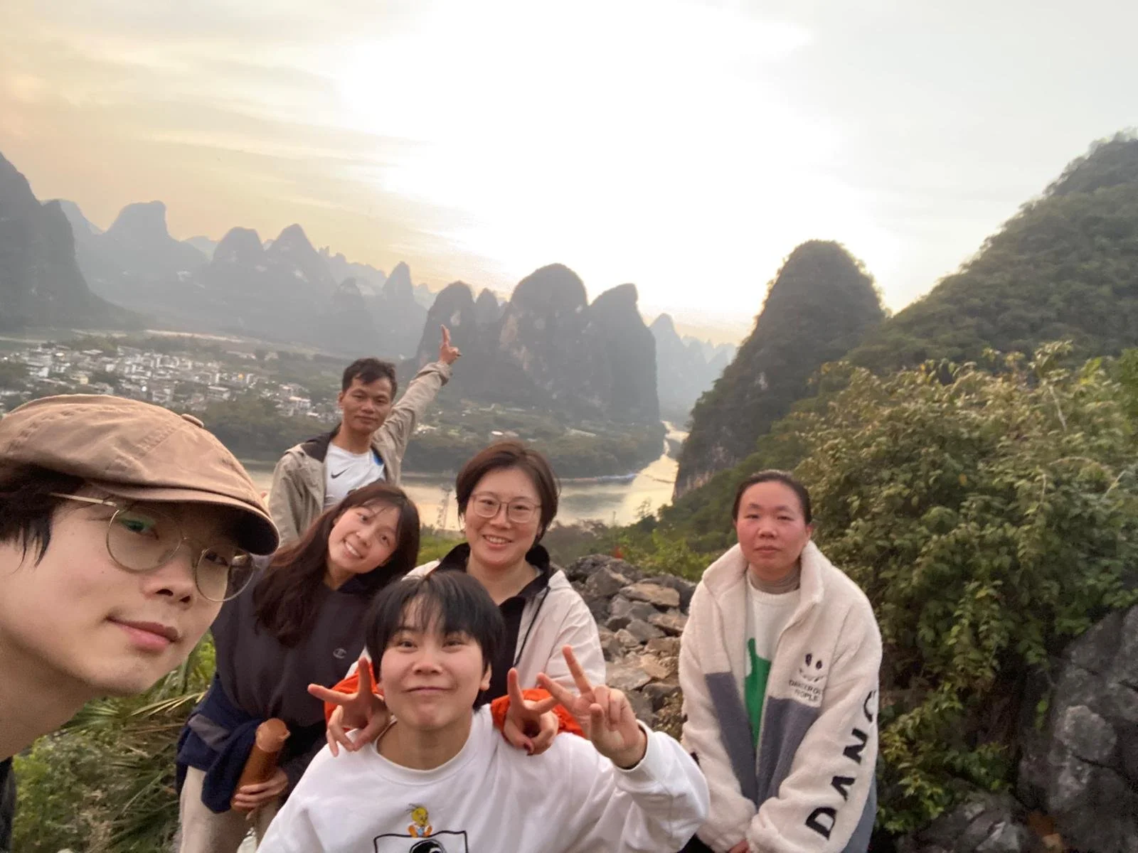 A group of six people taking a selfie with a scenic landscape of mountains, a river, and a village in the background during sunset.