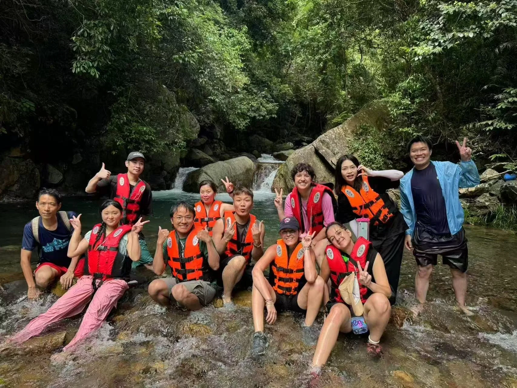 Group of people in a river surrounded by lush green trees, some wearing life jackets and smiling at the camera.