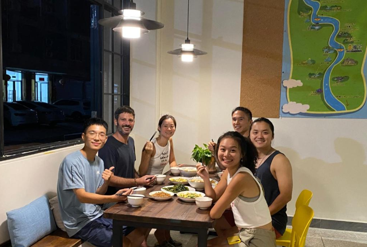 Group of six young adults enjoying a meal together at a wooden table indoors, with bowls of food and a potted plant, in a modern room with hanging lights and a large map on the wall.