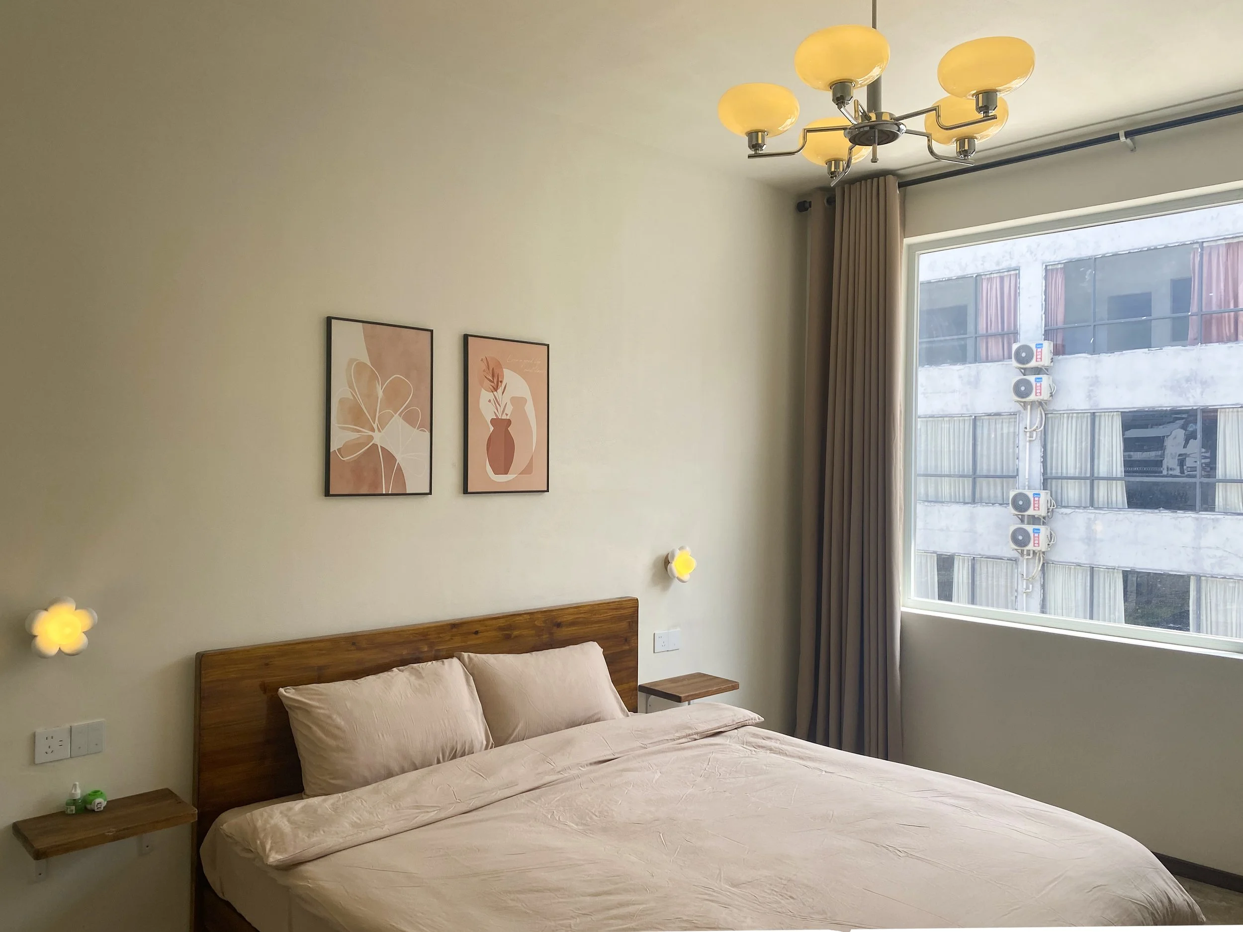 A bedroom with a wooden bed frame, beige bedding, and two pillows. There are two wall-mounted lights with yellow flower-shaped covers on either side of the bed. The wall above the bed has two framed art prints featuring minimalist floral designs in earthy tones. A large window with beige curtains shows a neighboring building with air conditioning units and gray concrete walls. A ceiling light fixture with multiple yellowish globes hangs from the ceiling.