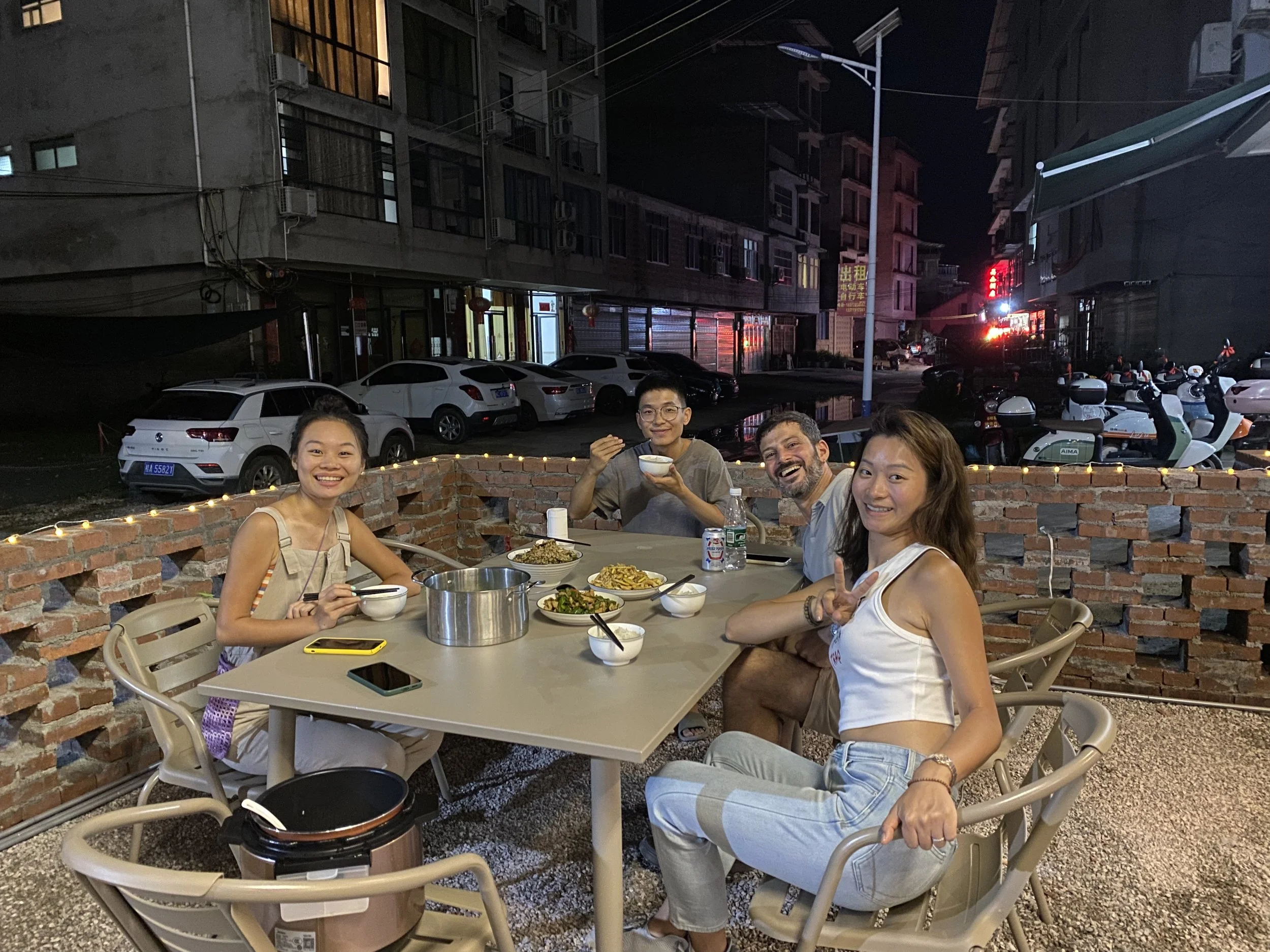 Four friends dining outdoors at night on a brick patio, enjoying food and drinks with city buildings and parked motorcycles in the background.