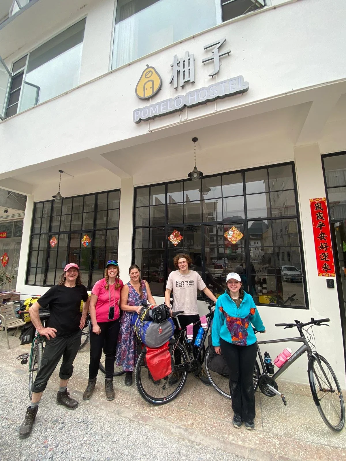 Group of five people with bicycles standing outside Pomelo Hostel, smiling, in front of the building with large windows and Chinese New Year decorations.