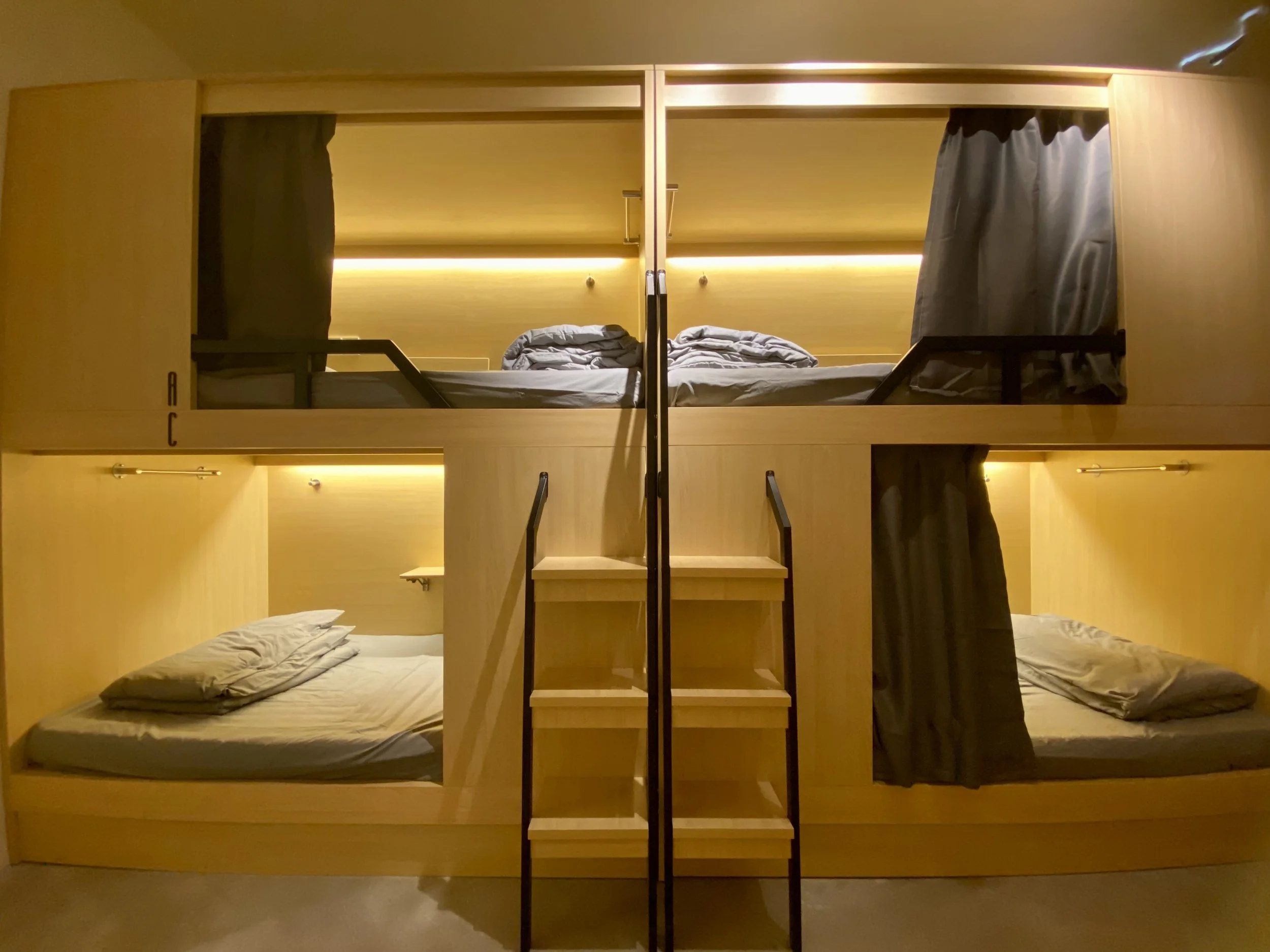 A modern wooden bunk bed structure with four beds, two on top and two below, in a room with warm lighting and gray bedding, accessed by a small staircase with black handrails.