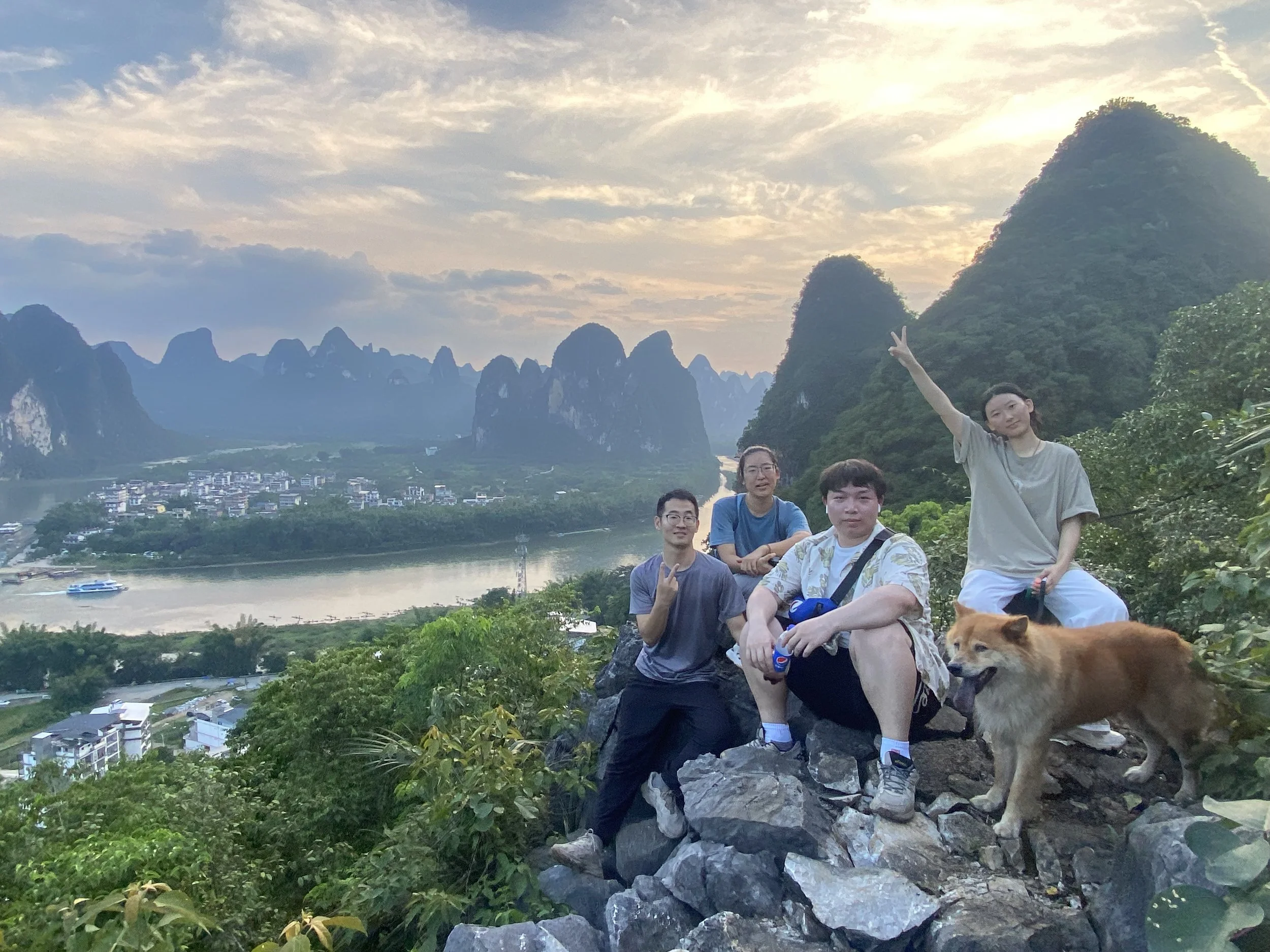 Four people and a dog on a rocky ledge overlooking a river and mountains at sunset.