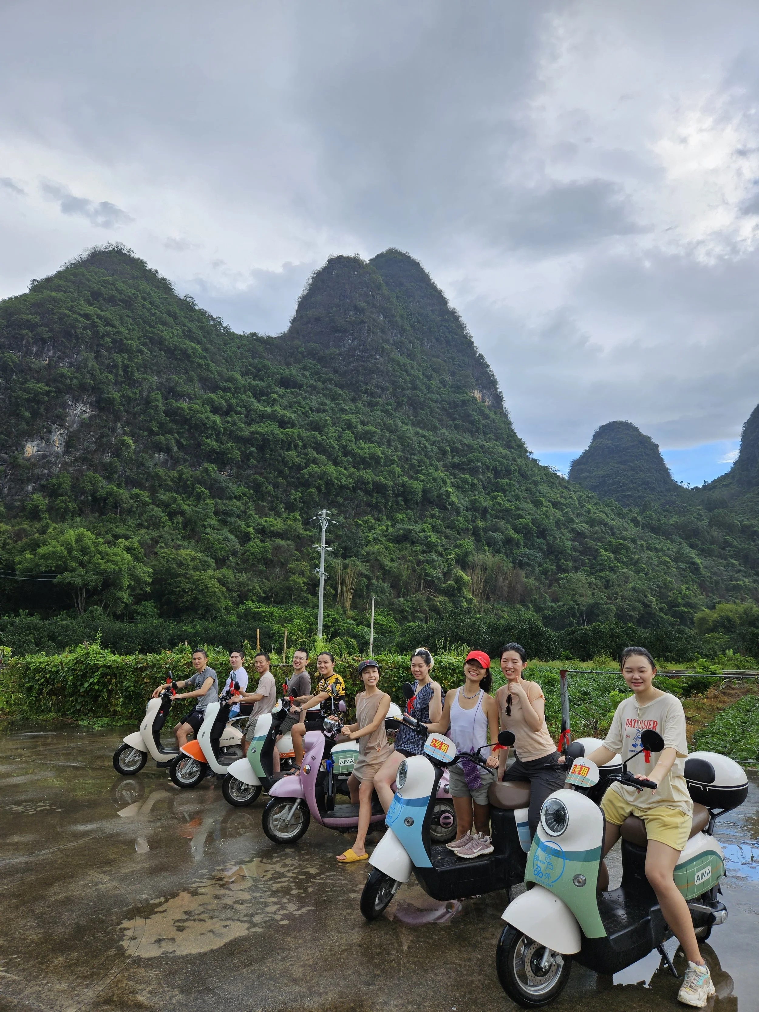 A group of people on scooters in a rainy outdoor setting with lush green mountains and cloudy sky in the background.