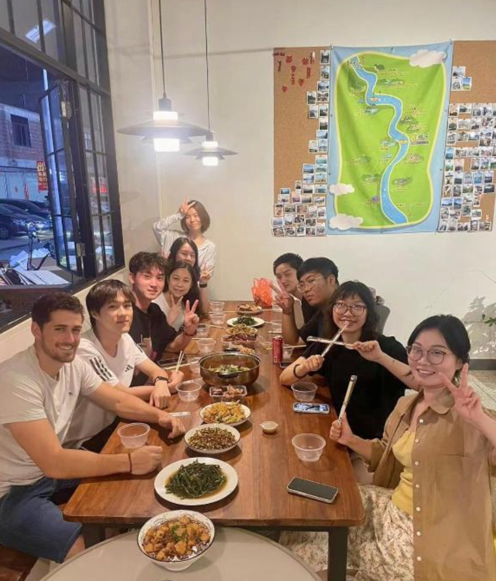 Group of ten people sitting around a table enjoying a meal at a restaurant, with dishes of Korean food, some smiling and making peace signs, in a cozy indoor setting with a large window and a map on the wall.
