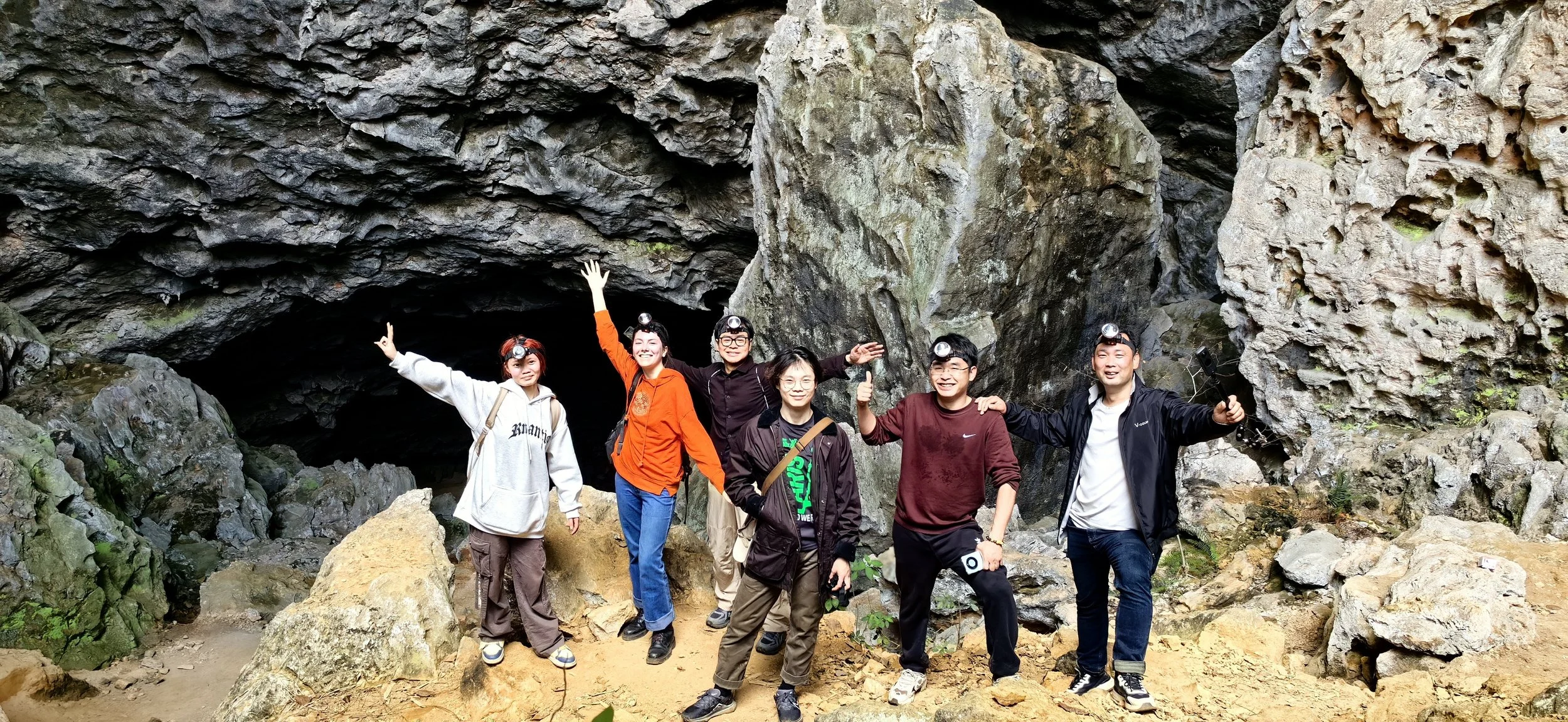 Group of six people hiking inside a rocky cave, wearing headlamps, some waving, smiling, and taking photos.