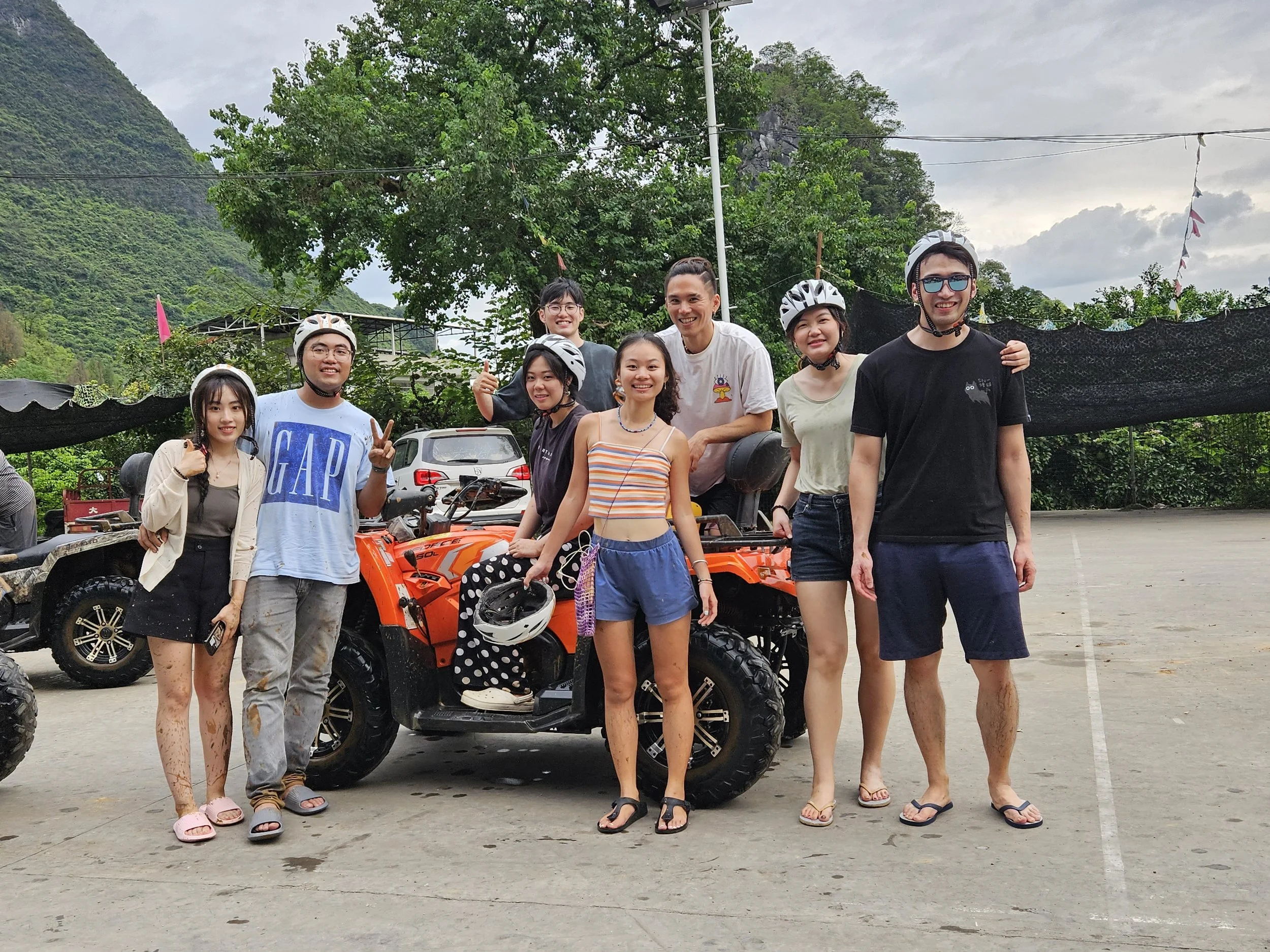 Group of nine young people, some wearing helmets, standing and sitting around an orange all-terrain vehicle (ATV) on a concrete parking lot outdoors. Some are smiling and posing for the camera with lush green mountains and trees in the background.