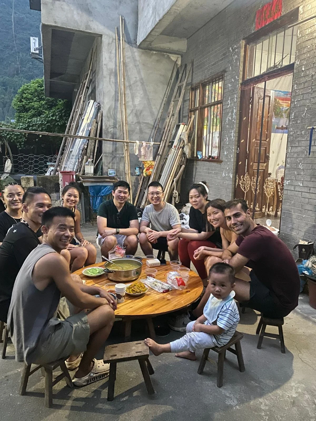 Group of people sitting around a low wooden table outdoors with food and drinks, smiling at the camera, with a small child sitting on a stool in front.