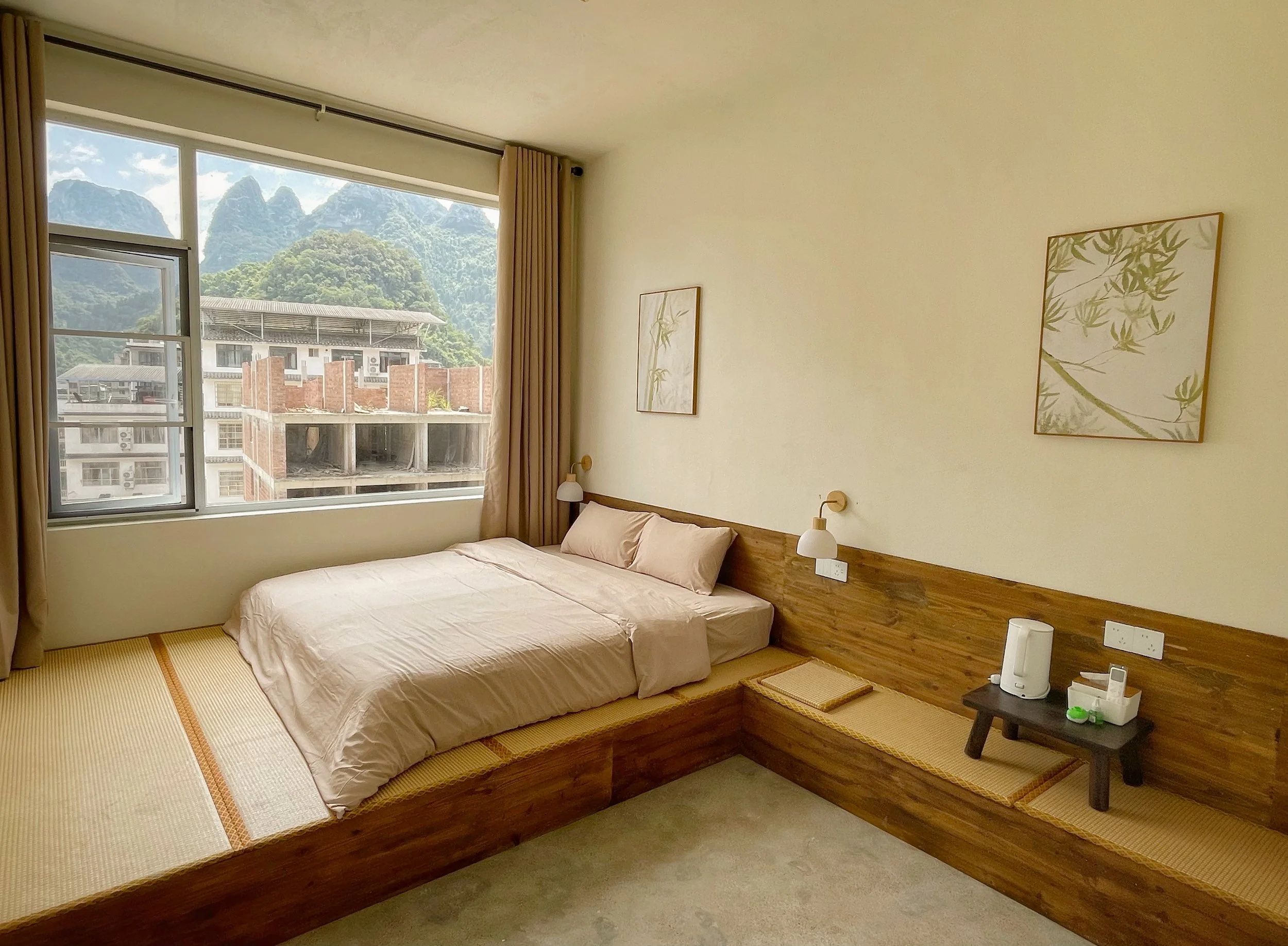 Hotel room with a large window showing mountains and buildings outside, a bed with beige linens, bedside lamps, and minimalist wall art.