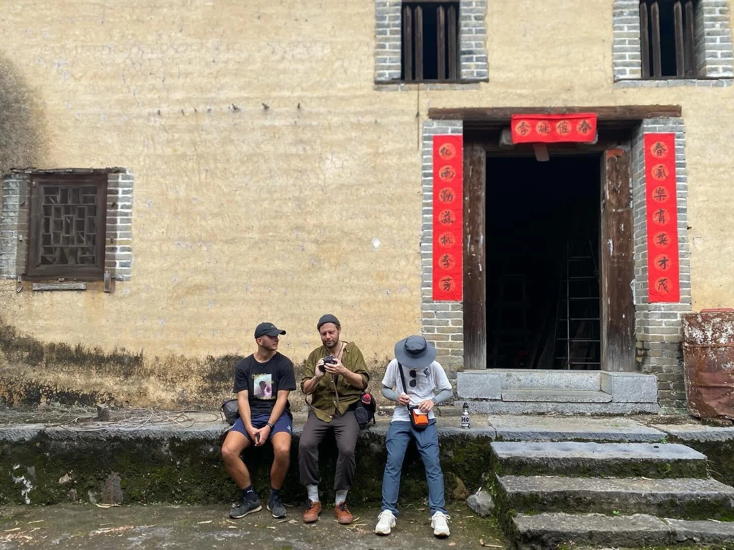 Three people sitting on a ledge in front of an old yellow brick building with red Chinese banners beside the door.