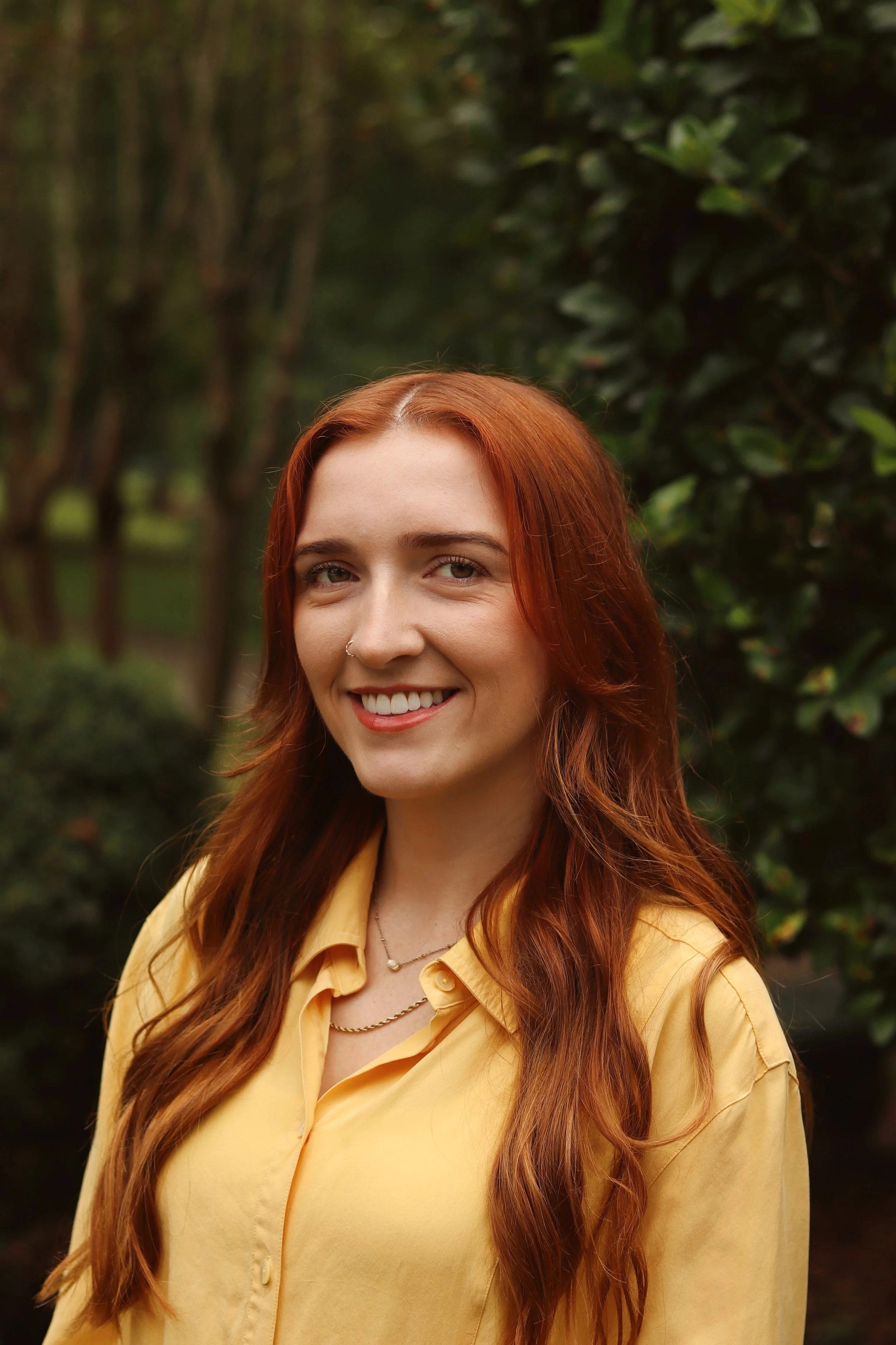 Young woman with long, wavy red hair smiling outdoors, wearing a yellow blouse and a necklace, standing in front of green foliage.