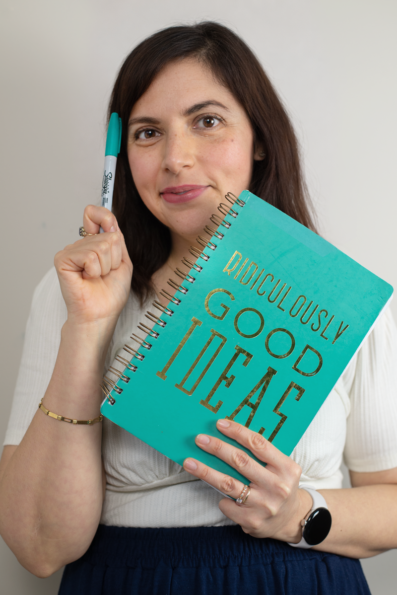 Ana Miller, woman with dark brown hair holding a teal notebook with gold lettering that says 'Ridiculously Good Ideas.' She is holding a teal marker near her temple and smiling at the camera.