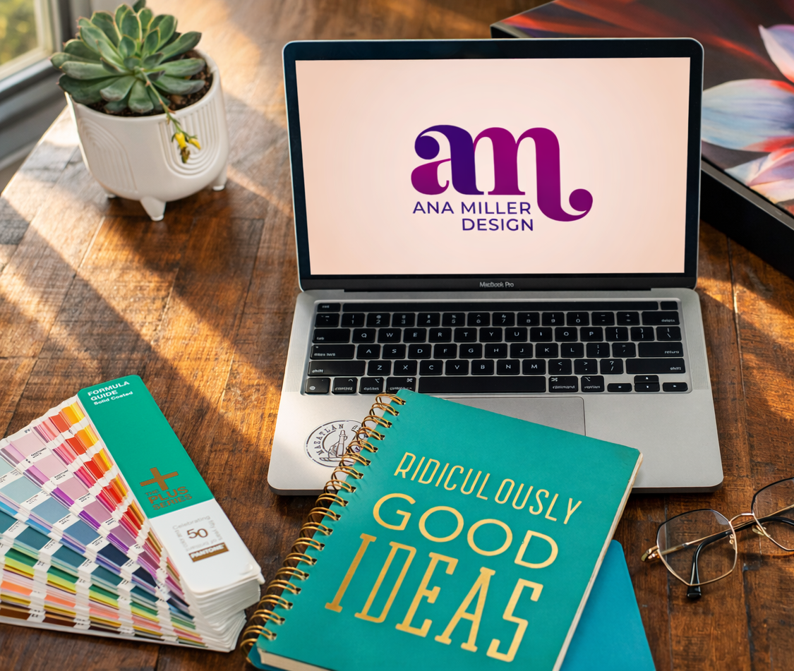 Open MacBook Pro displaying a logo for Ana Miller Design, a color palette sample book, a spiral notebook titled "Ridiculously Good Ideas," a pair of glasses, and a small potted succulent on a wooden desk with sunlight streaming in.