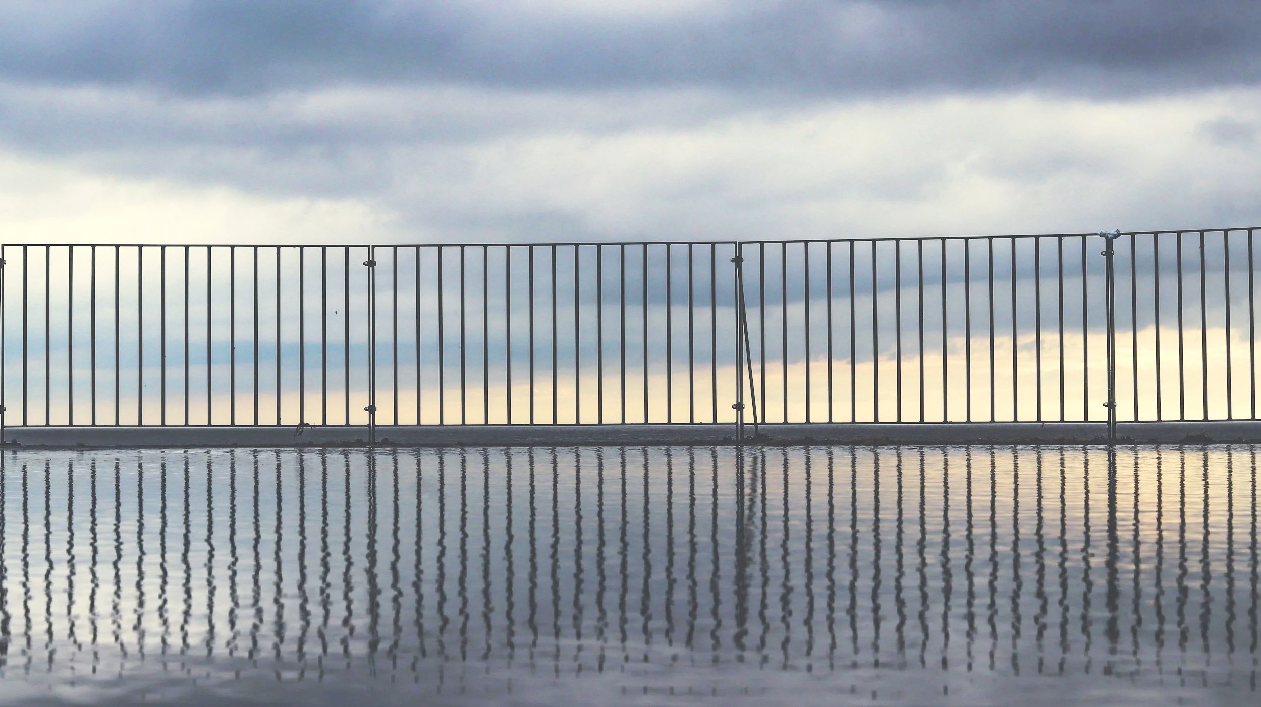 A metal safety fence on a bridge over water, with the water reflecting the fence and a cloudy sky in the background.