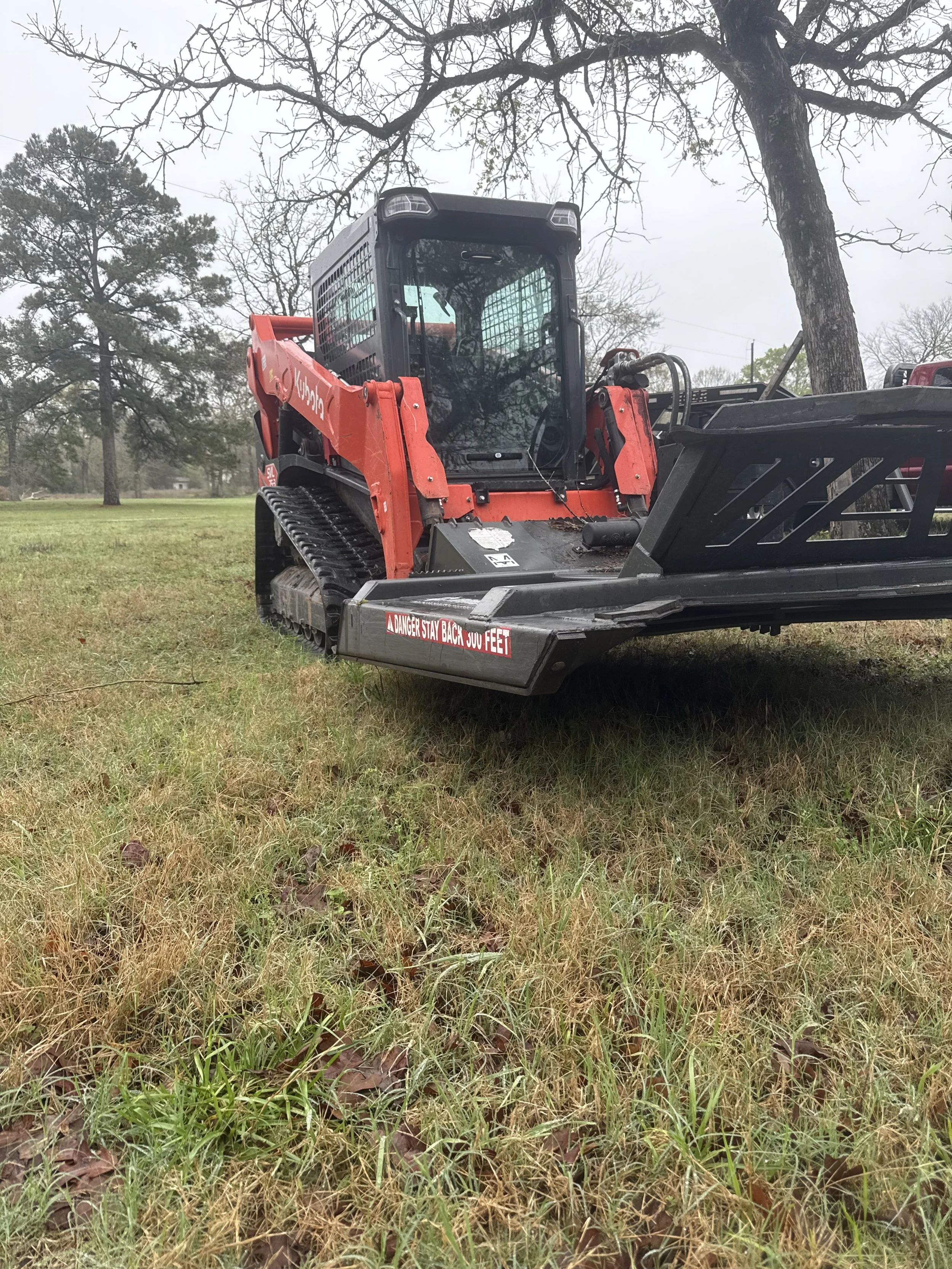 A red compact track loader with a black attachment parked on a grassy field, with trees in the background.