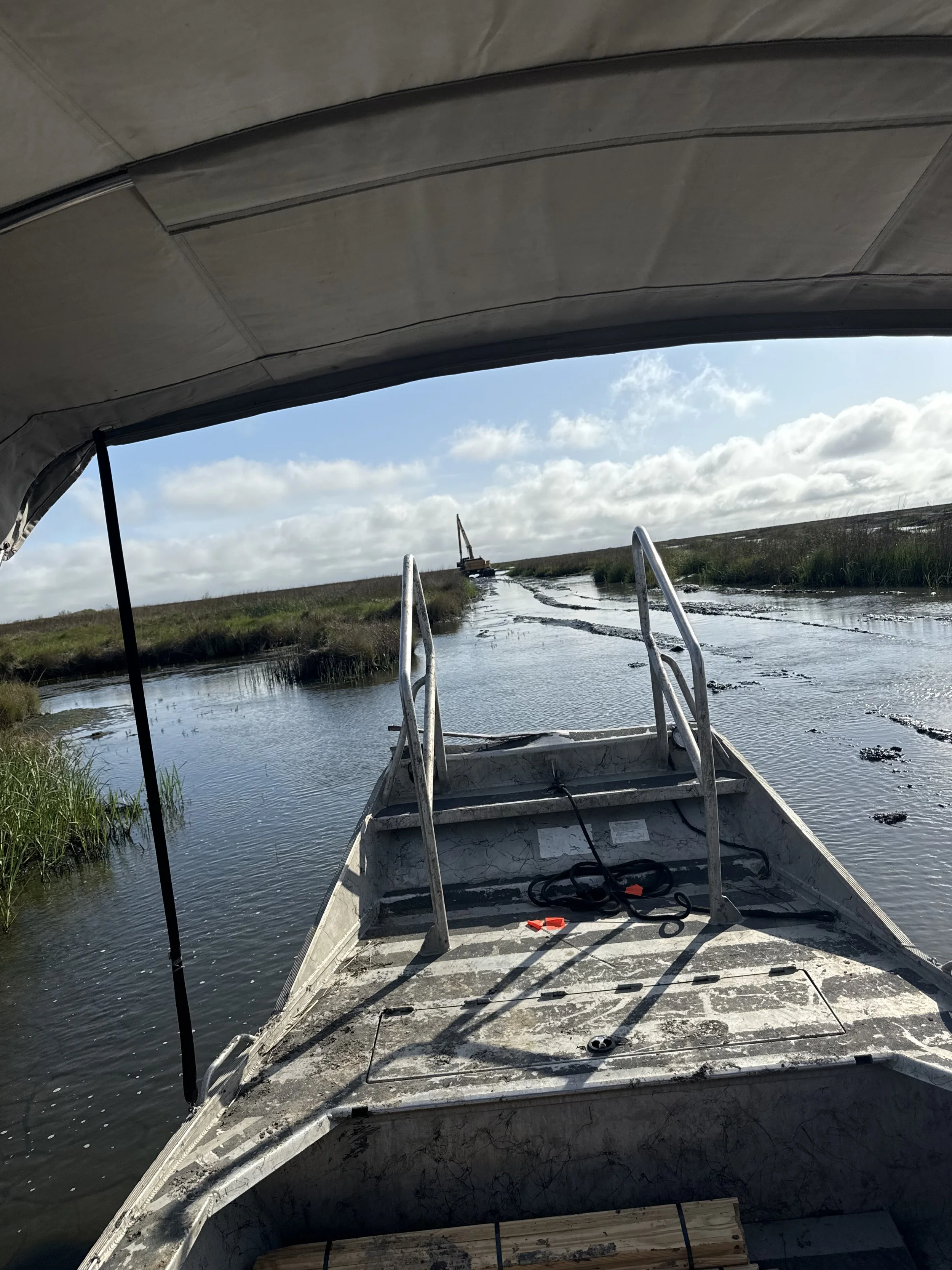 View from a boat heading down a narrow waterway with grassy banks, under a partly cloudy sky, with a crane visible in the distance.