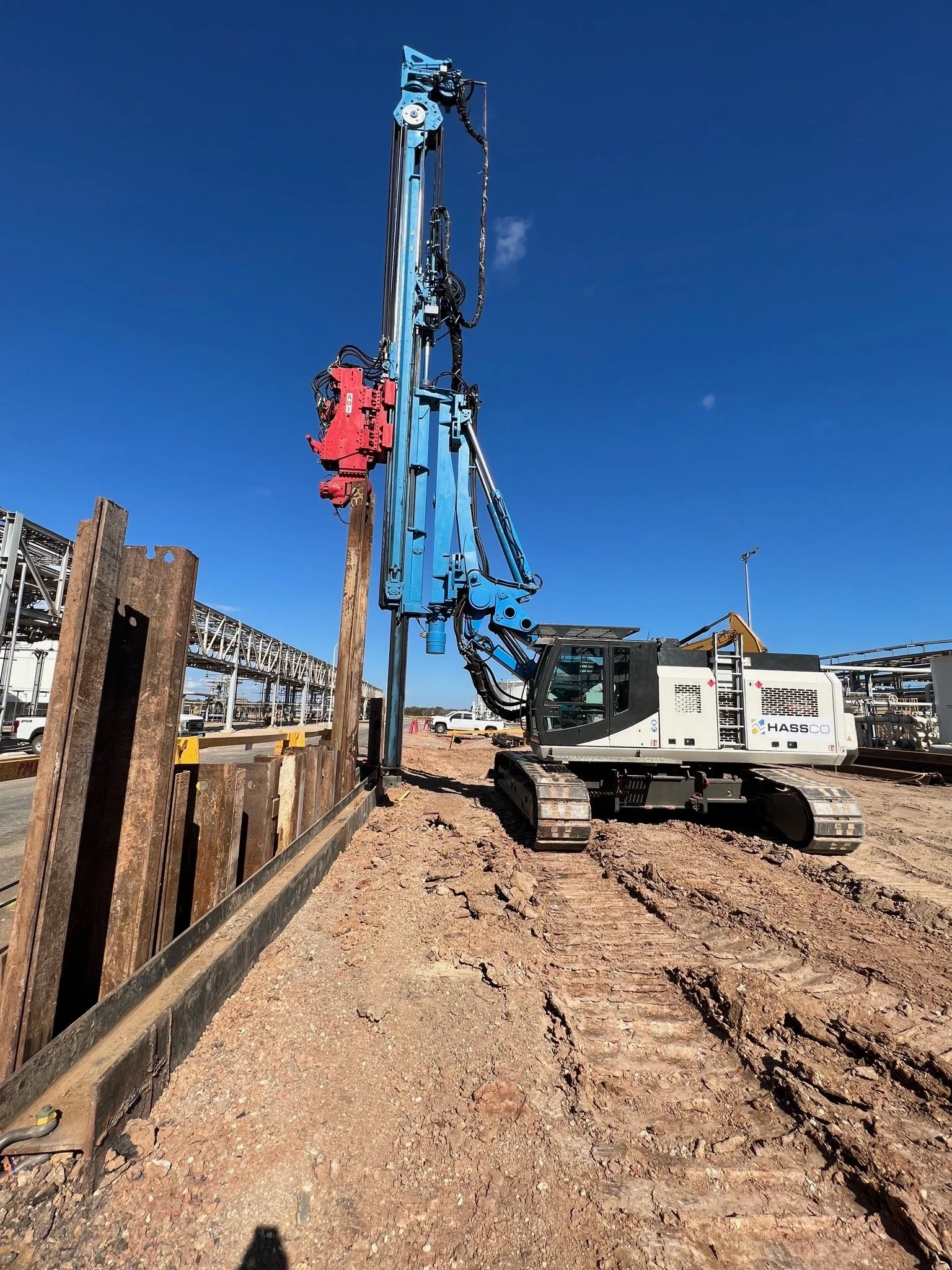 Construction excavator with a large blue boom arm operating at a construction site, with wooden and metal barriers and dirt ground under a clear blue sky.