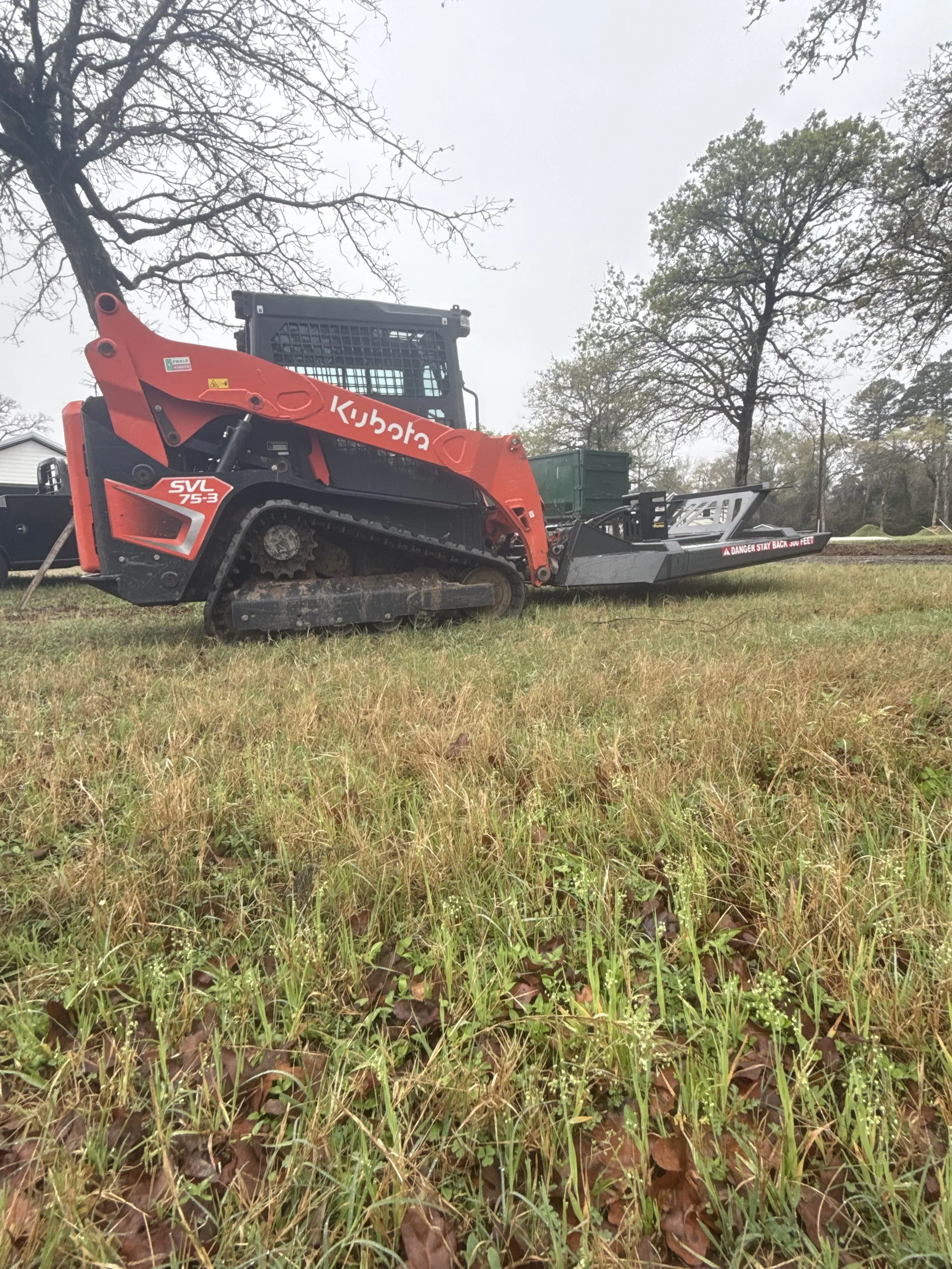 A Kubota SVL 75-3 compact track loader parked on a grassy field with trees in the background.