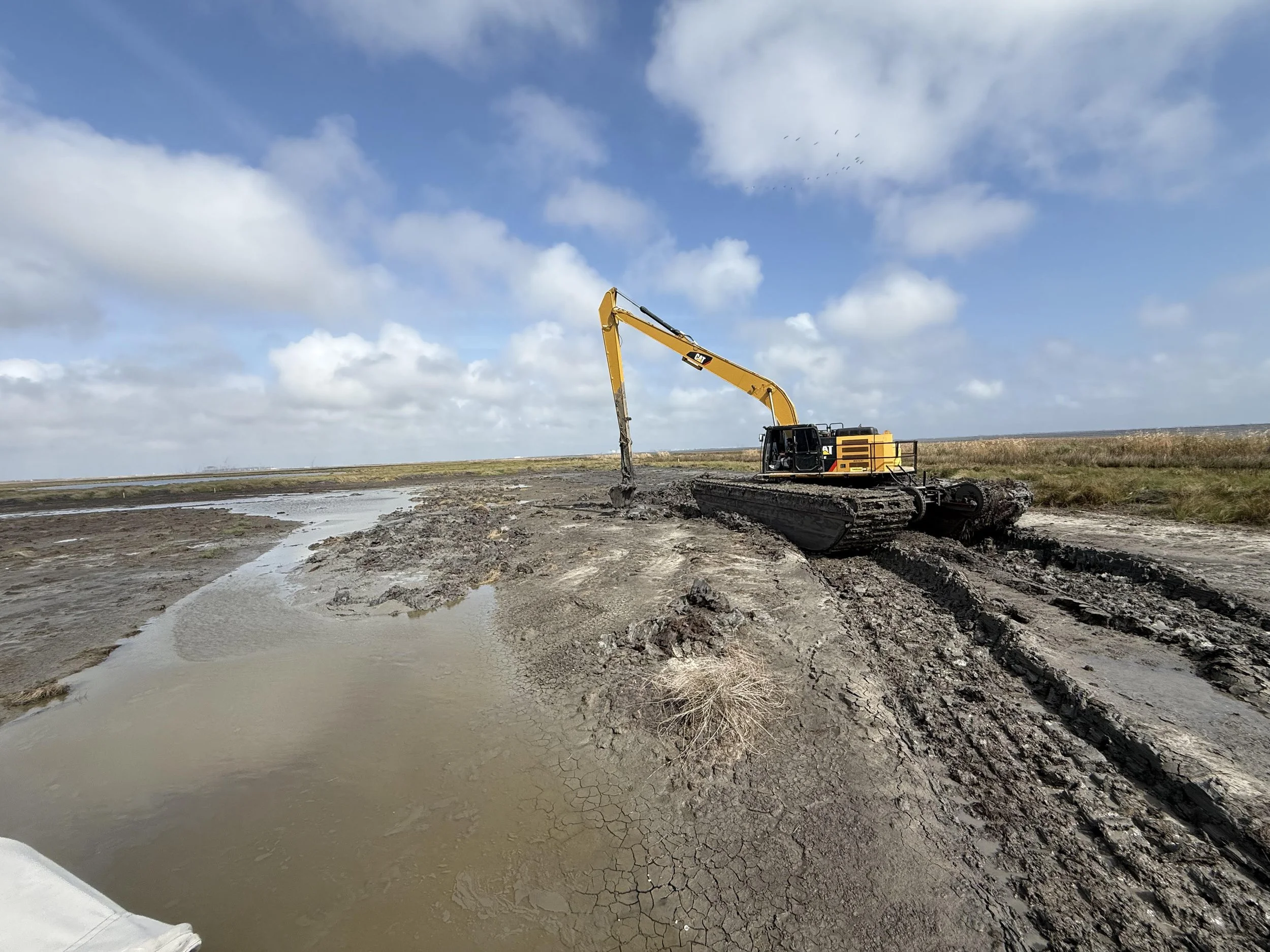 A yellow excavator working on a dry, cracked landscape with a small water channel and grassy terrain under a partly cloudy sky.