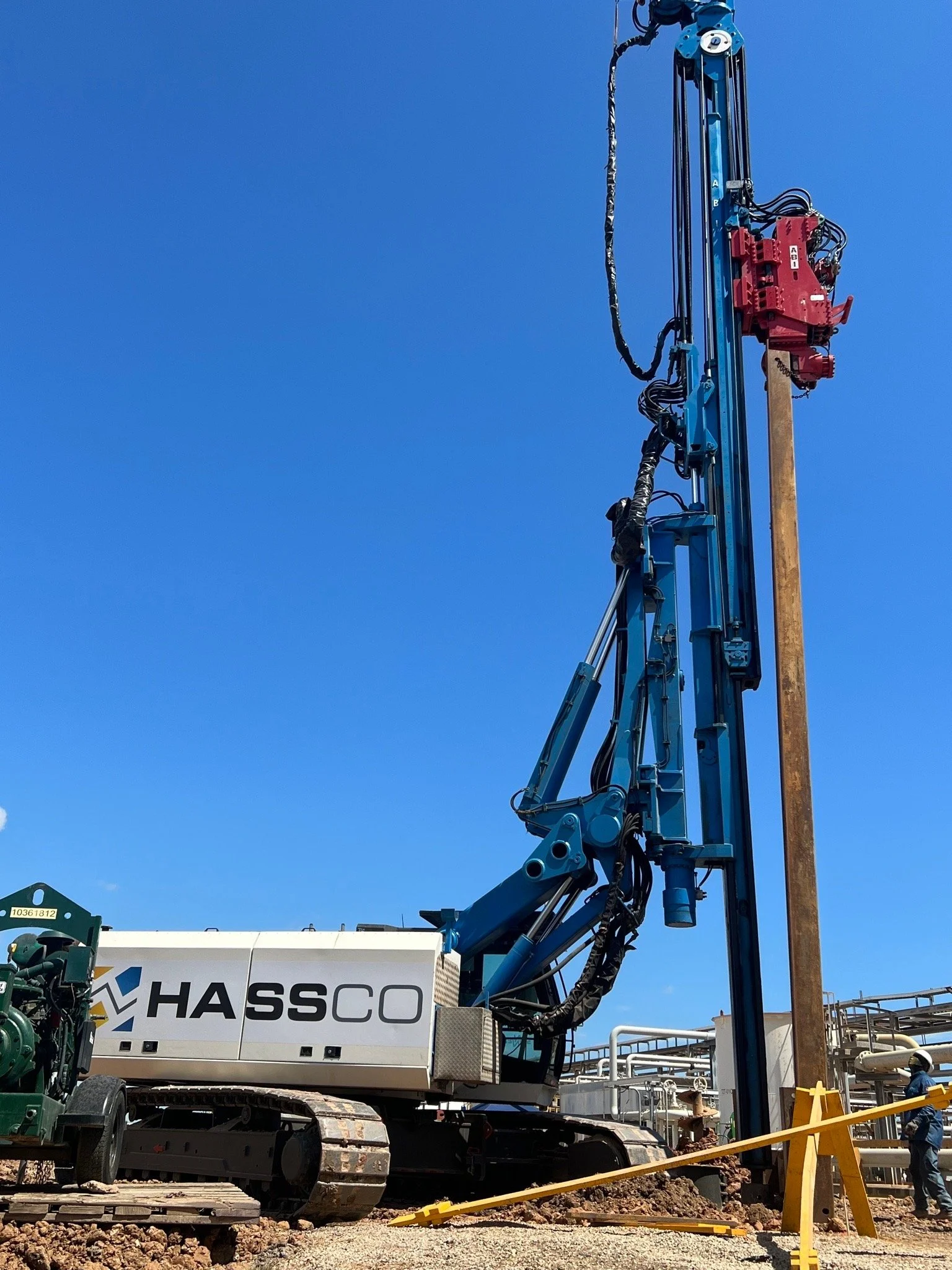 Large blue and red drilling machine at an industrial site with a worker in safety gear nearby.